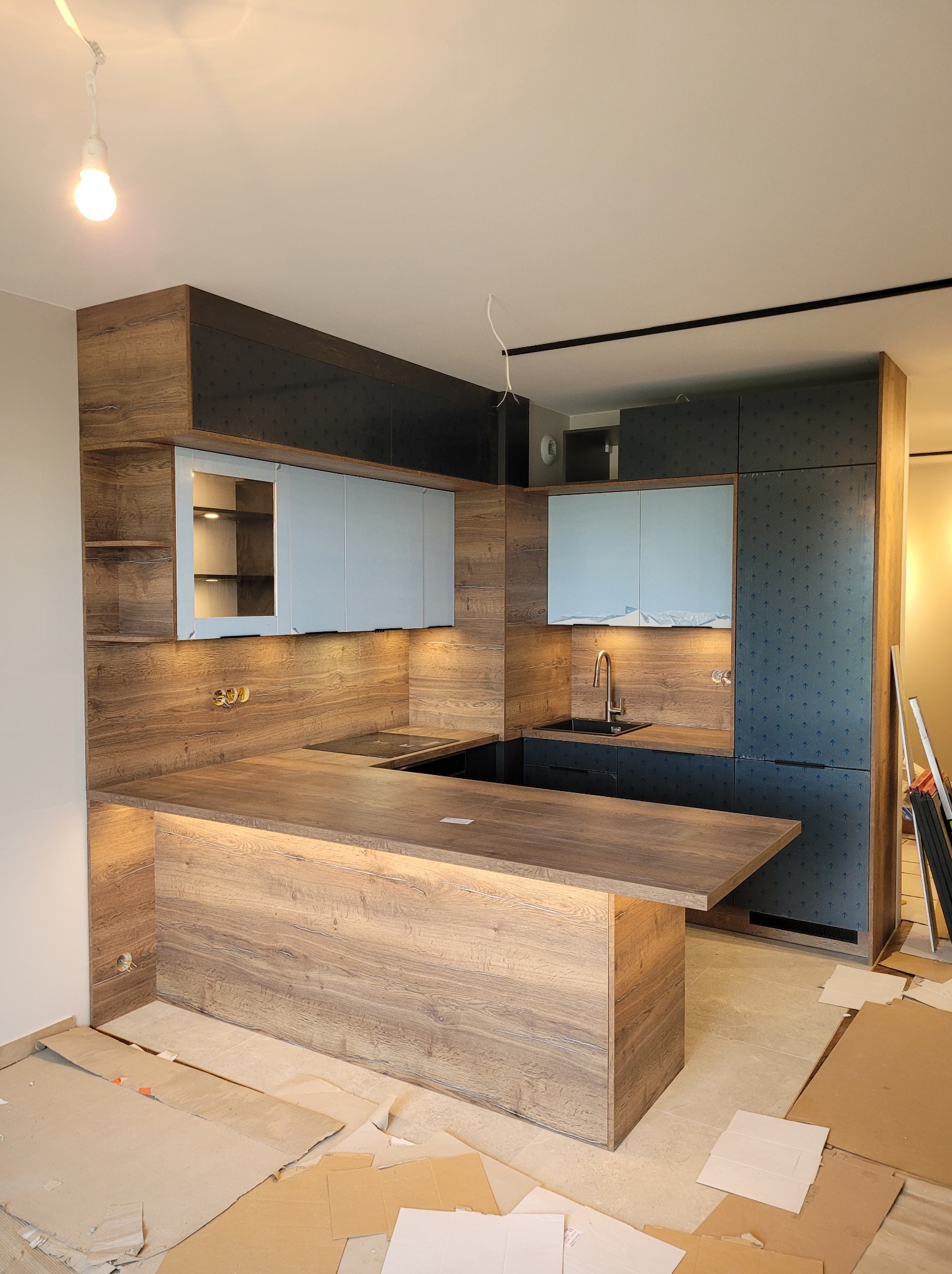 Nowoczesna kuchnia w trakcie montażu: drewniane fronty szafek, podświetlany blat wyspy, niebieskie górne szafki, widoczne elementy instalacji elektrycznej i kartony zabezpieczające podłogę.