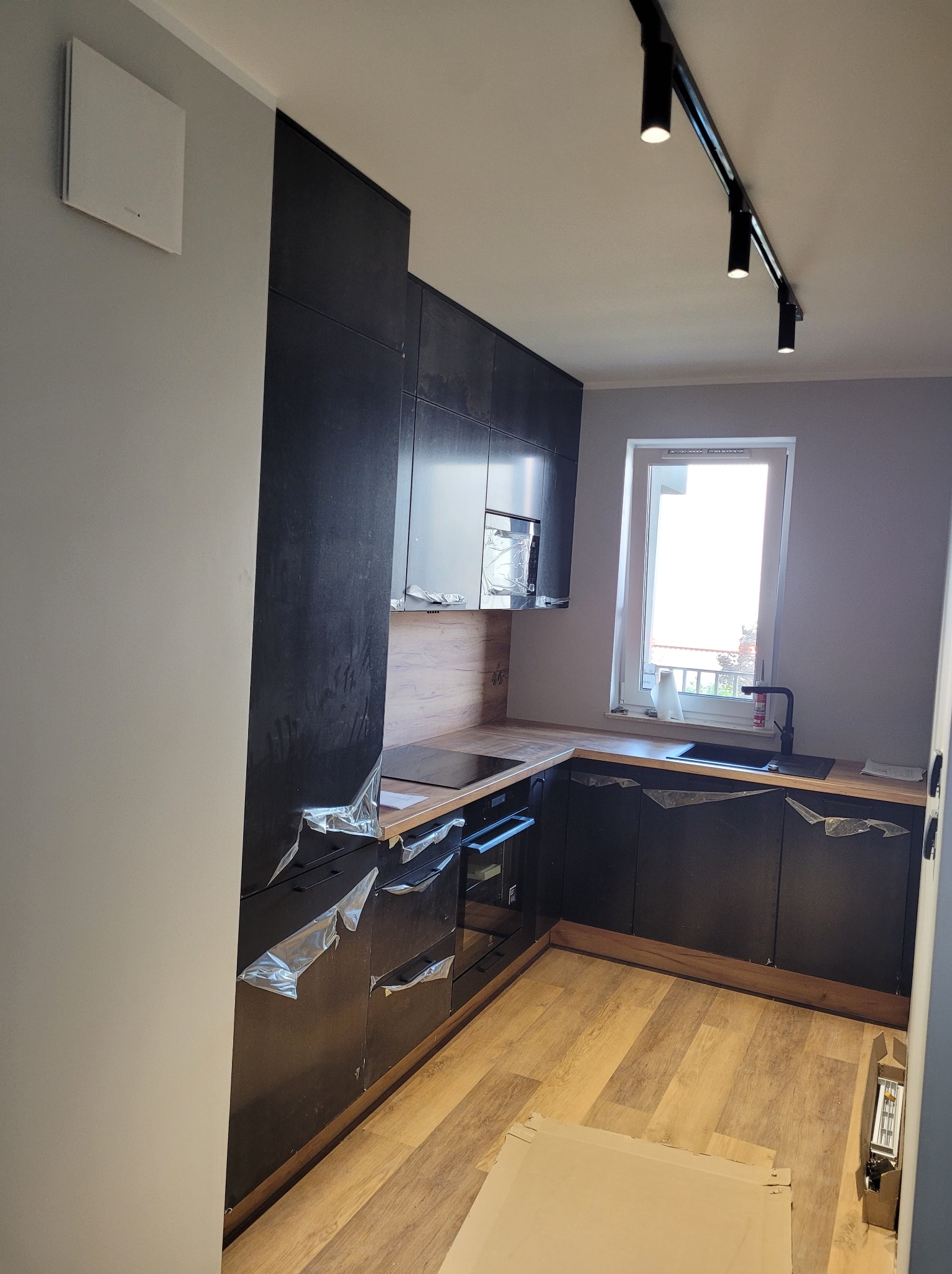 Ciemna, nowoczesna kuchnia z drewnianymi blatami, częściowo zabezpieczona folią ochronną, z widocznym oświetleniem punktowym na suficie i jasnym oknem w tle.