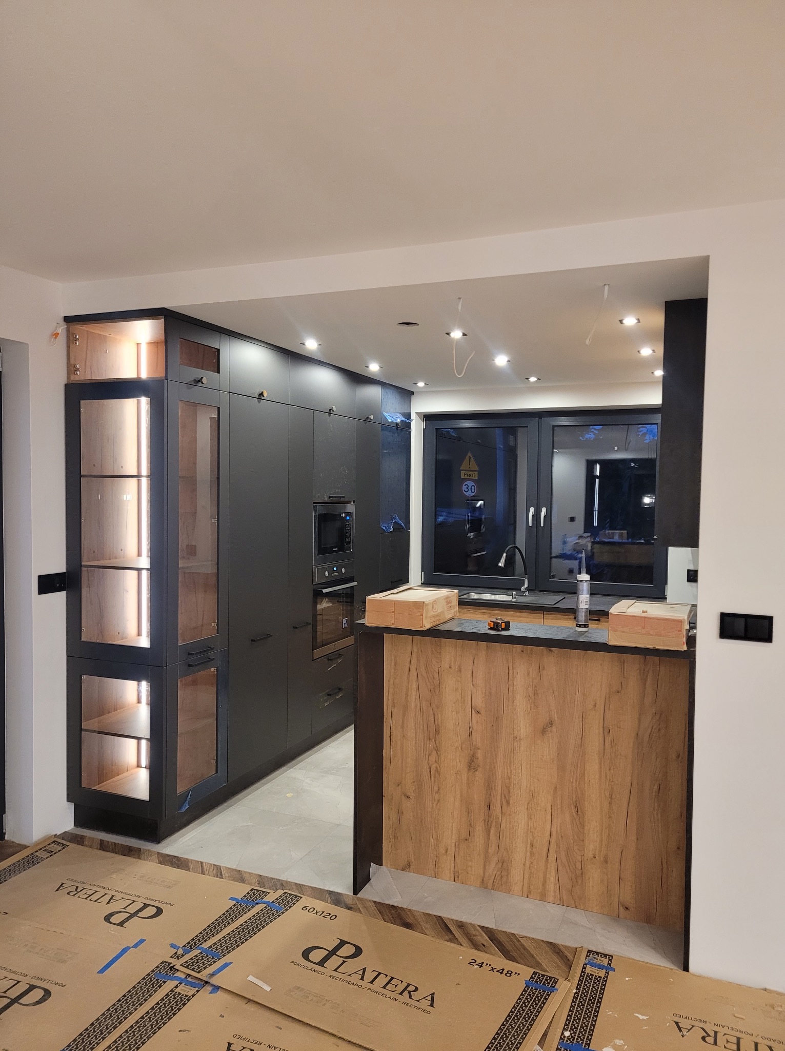 Ciemna, nowoczesna kuchnia z oświetleniem LED w witrynach, widok na blat z drewnianym wykończeniem i okno z widokiem na zewnątrz, na podłodze kartony z płytkami.