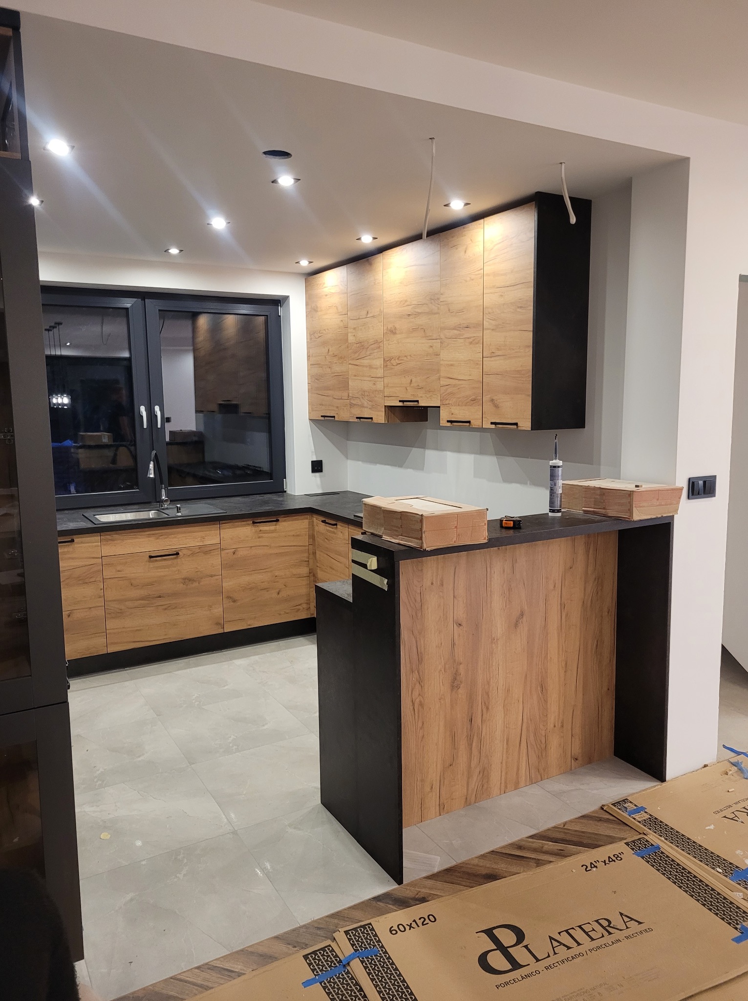 Montaż kuchni z drewnopodobnymi frontami i czarnymi elementami, widoczne kartony z płytkami podłogowymi i silikon na blacie, oświetlenie sufitowe.