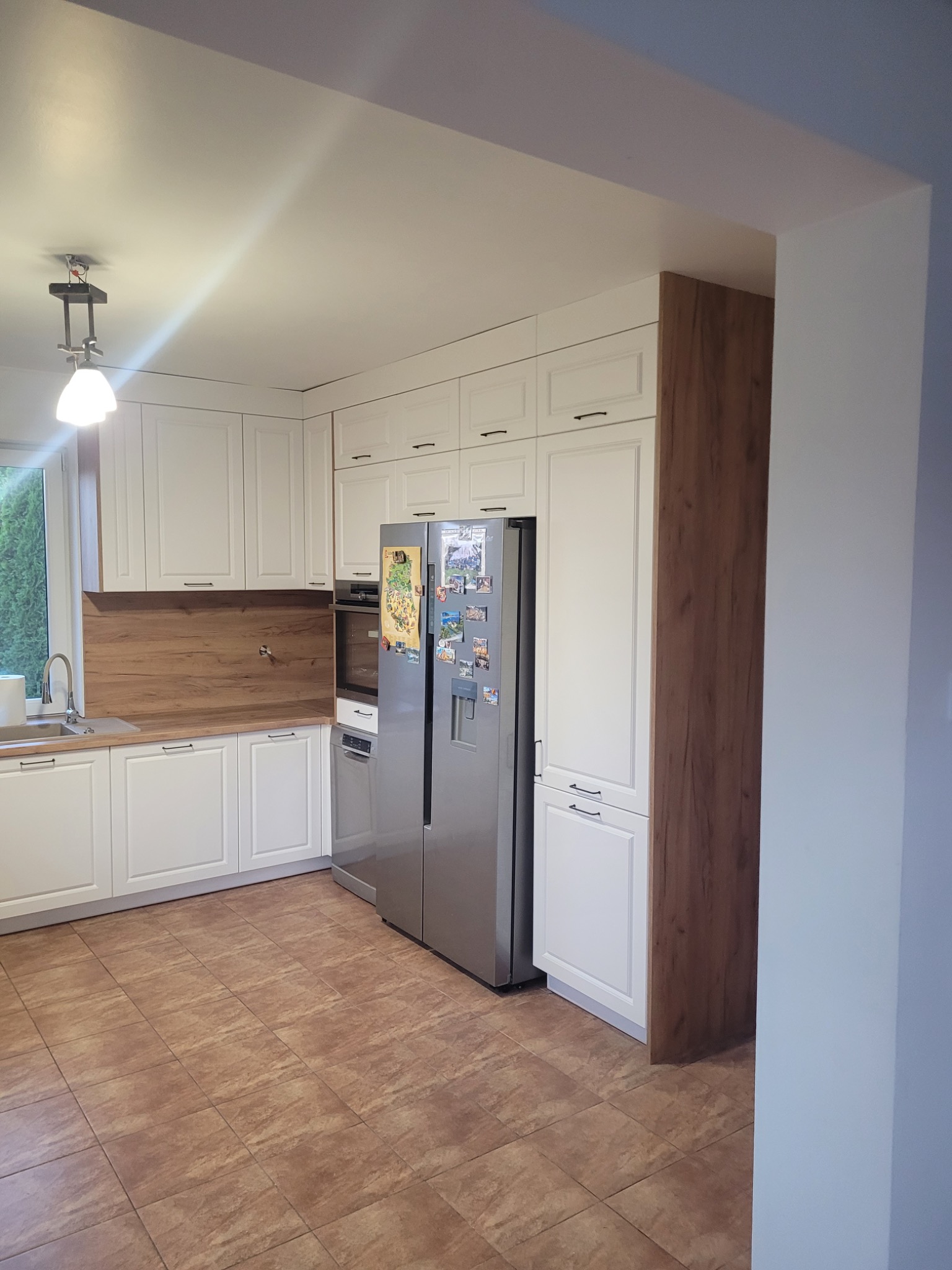 Jasna kuchnia z białymi frontami szafek, drewnianym blatem i brązowymi płytkami podłogowymi. Duża, dwudrzwiowa lodówka ze stali nierdzewnej z magnesami.