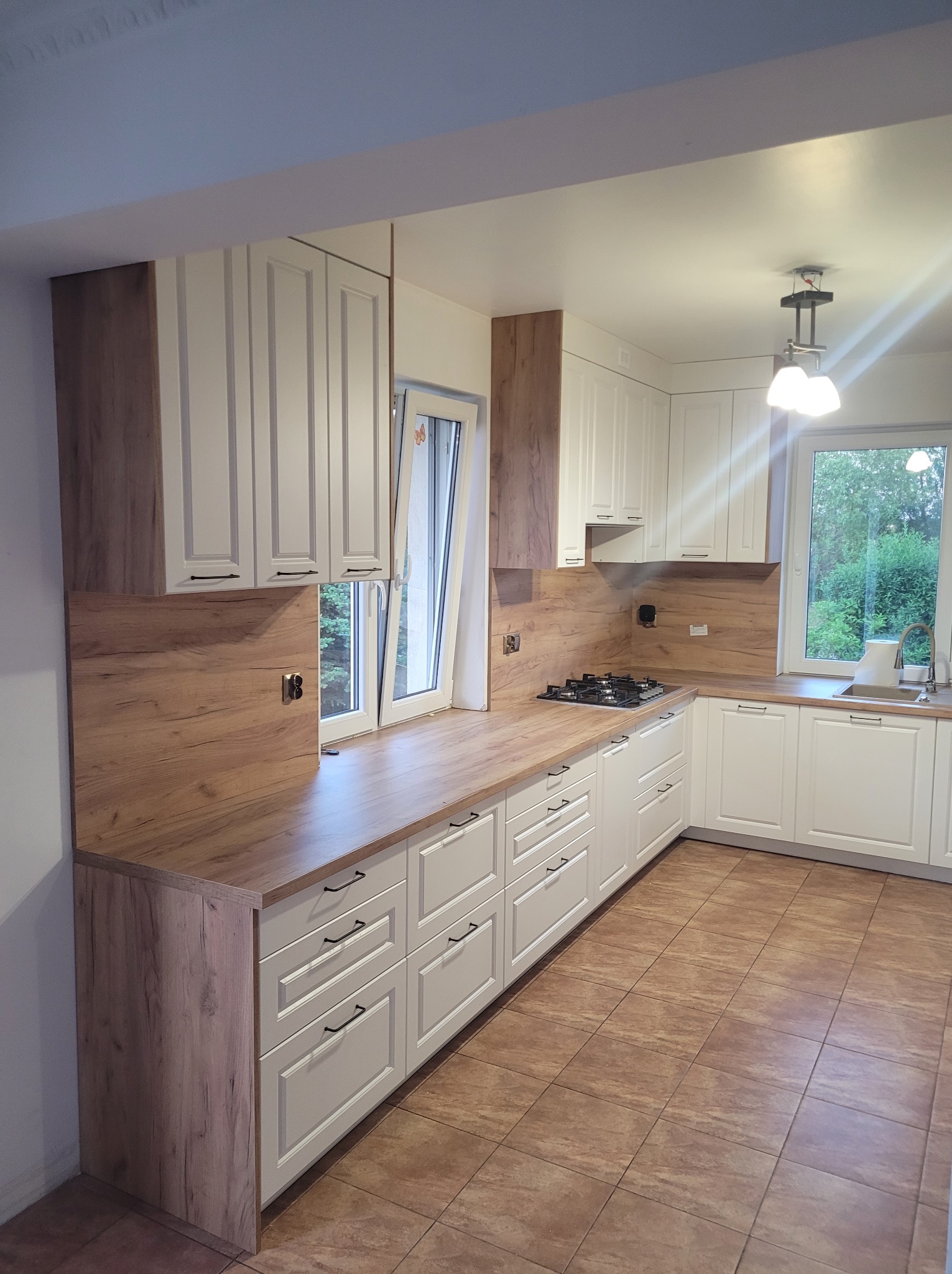 Jasna kuchnia z drewnianymi akcentami: kremowe szafki z frezowanymi frontami, drewniany blat i panel ścienny, brązowe płytki podłogowe, okno z widokiem na zieleń.