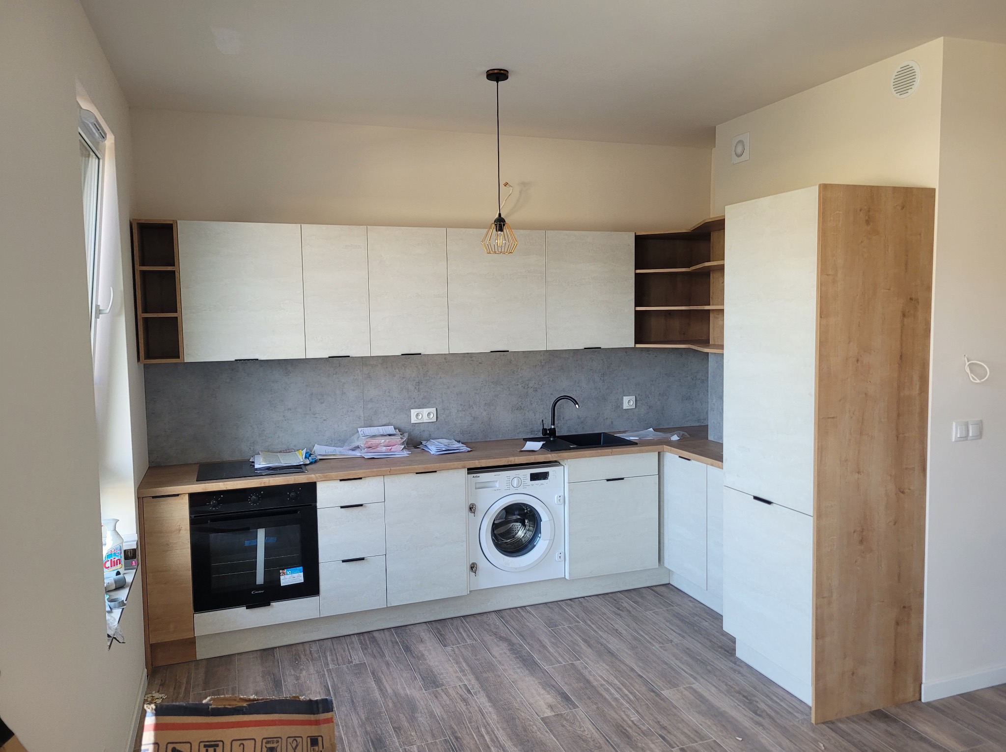 Jasna kuchnia z drewnianymi akcentami, zabudowana pralka, szare płytki na ścianie, lampa wisząca z metalowym ażurem, dokumenty i akcesoria na blacie.