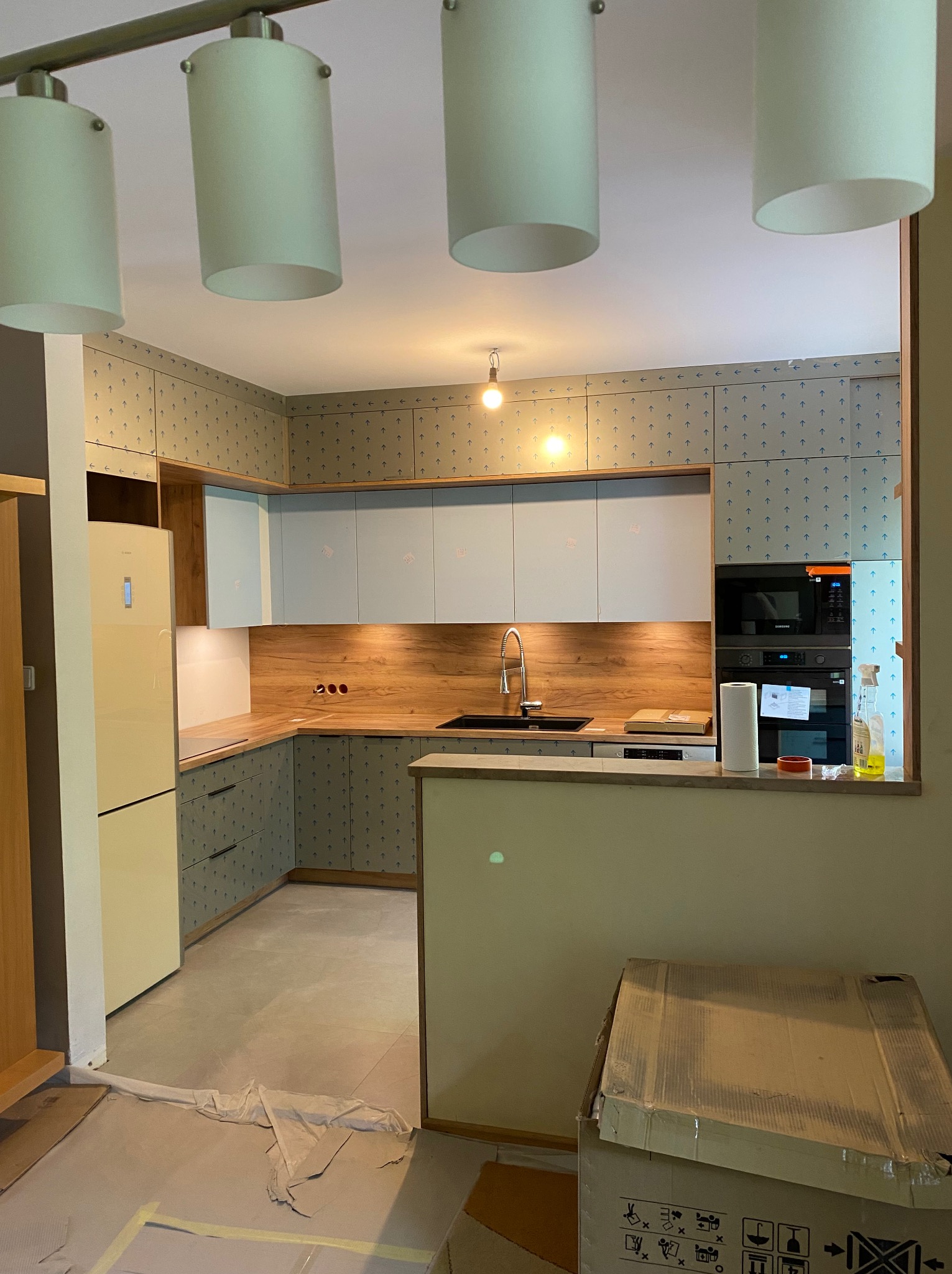 Nowoczesna kuchnia w trakcie montażu: jasne fronty szafek, drewniany blat, ściany wyłożone płytami z wzorem strzałek, podłoga zabezpieczona folią malarską, widoczne kartony z wyposażeniem.
