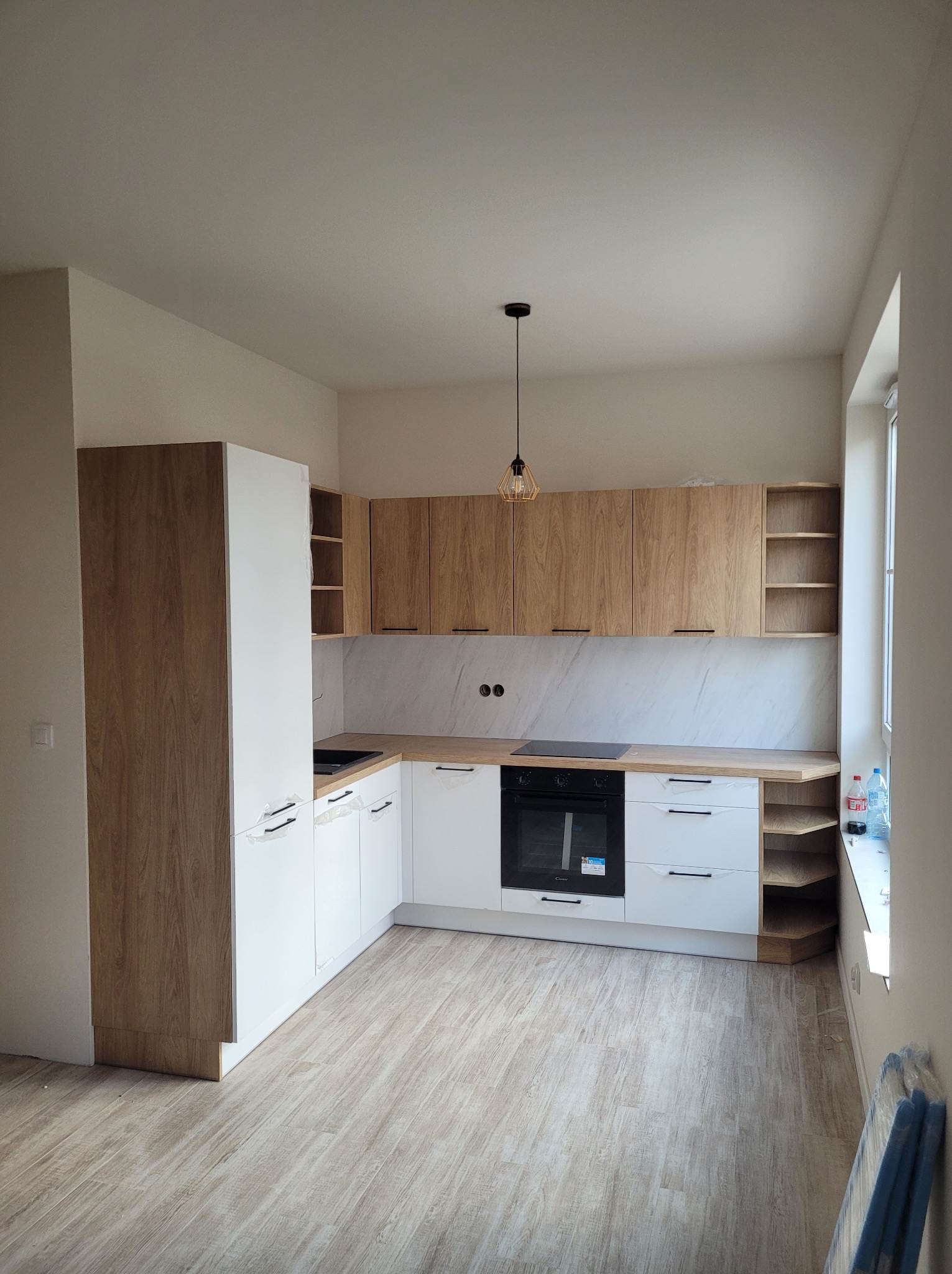 Nowoczesna kuchnia z frontami z jasnego drewna i białymi szafkami, marmurowy backsplash, drewniana podłoga, minimalistyczna lampa wisząca, częściowo jeszcze zabezpieczone folią uchwyty szafek.