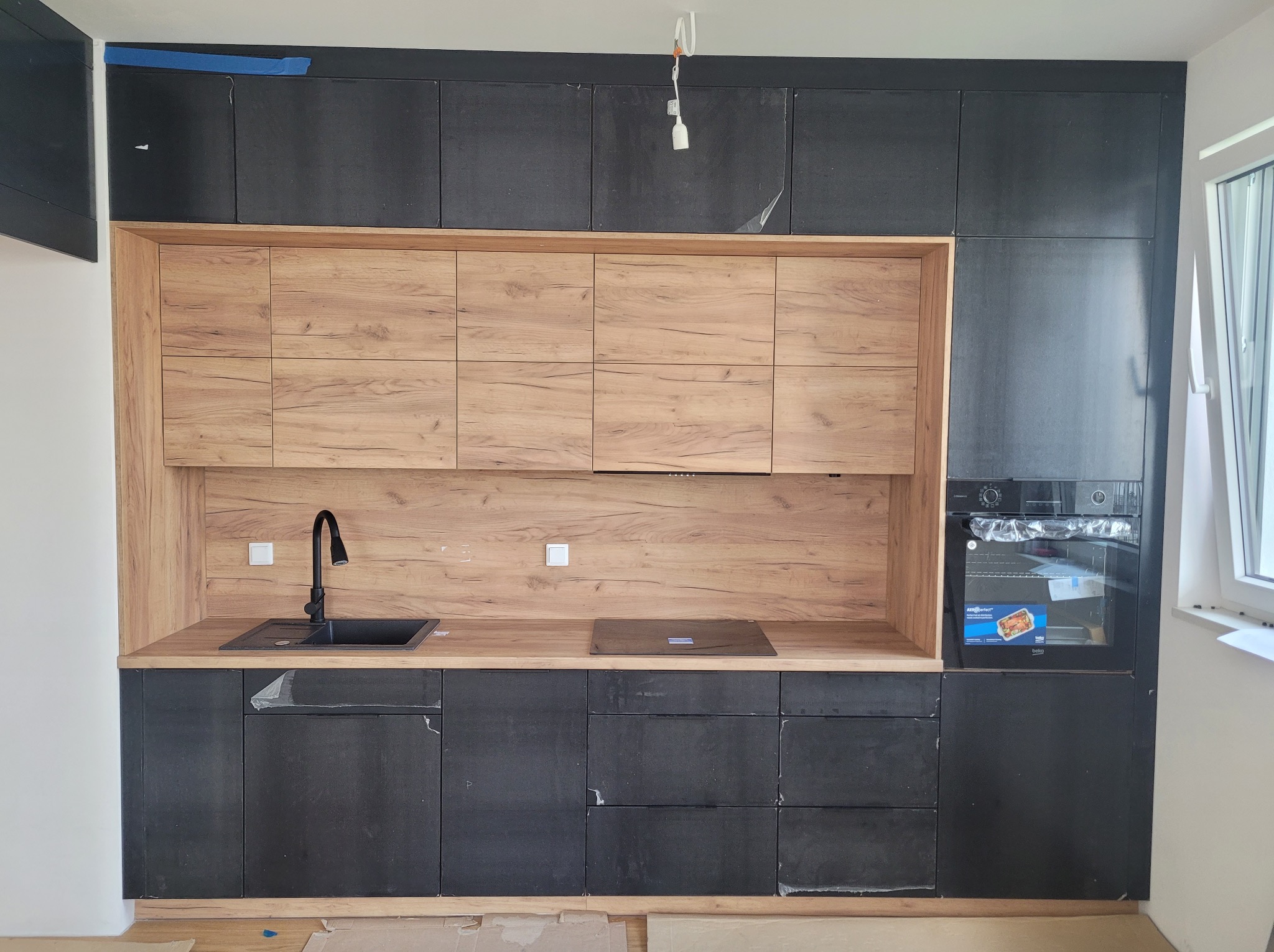 Nowoczesna zabudowa kuchenna z czarnymi szafkami dolnymi i górnymi oraz drewnopodobnymi szafkami wiszącymi, czarny zlew i płyta indukcyjna, piekarnik w zabudowie, widoczne folie ochronne na frontach.