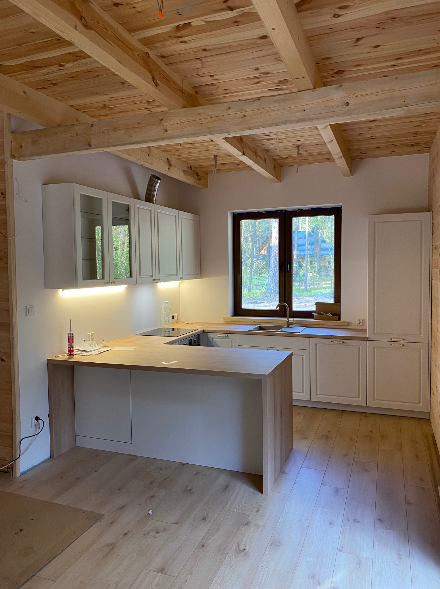 Kuchnia w trakcie montażu: białe szafki z drewnianymi blatami, wyspa kuchenna, drewniany sufit z belkami, widok na las za oknem, elementy wykończeniowe i narzędzia na blacie.