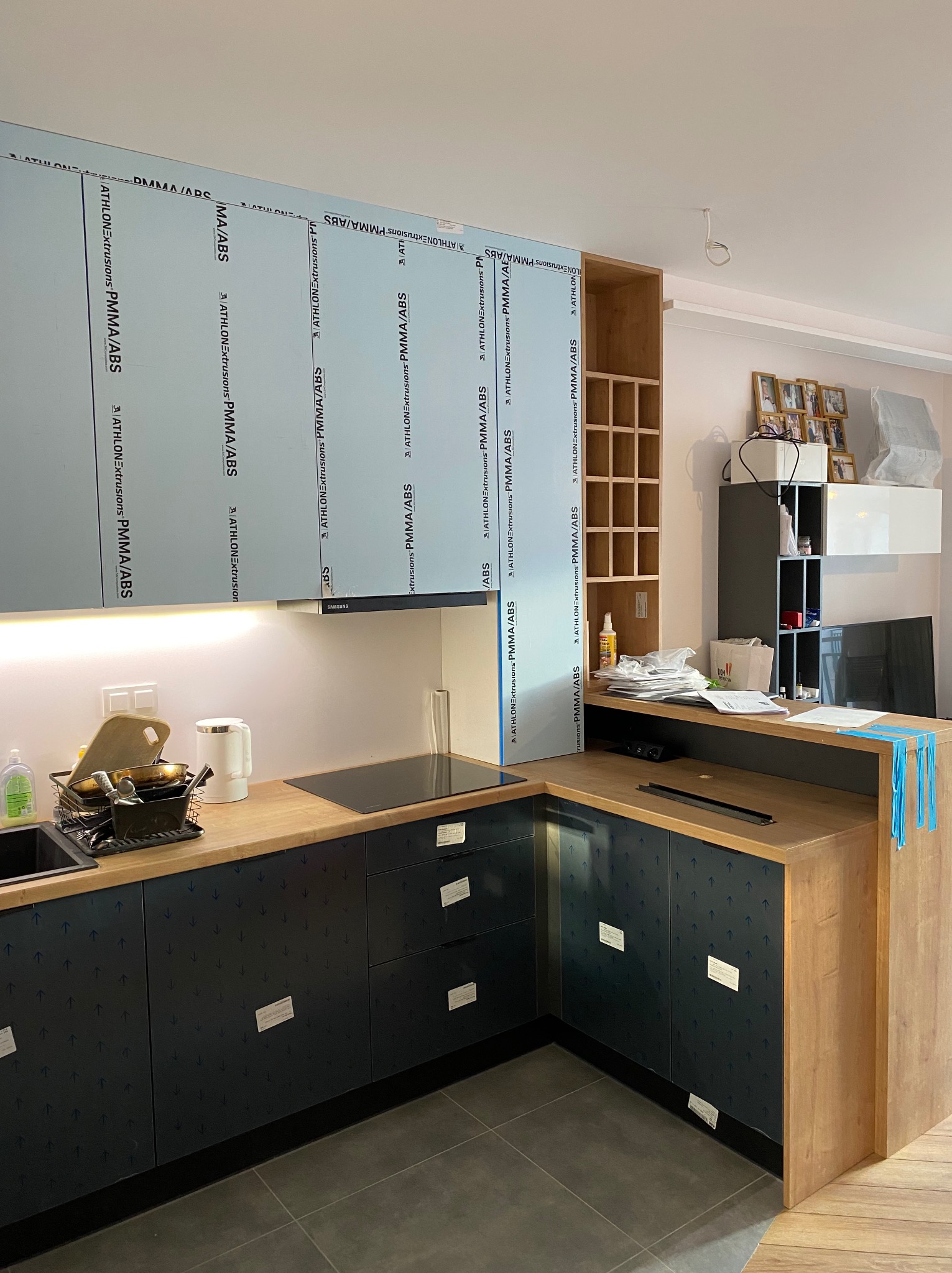 Montaż kuchni w Łochowie: widoczne zabezpieczone folią fronty szafek z wzorem strzałek, drewniany blat i ścianka, płyta indukcyjna, okap i częściowo zdemontowane płyty ochronne na ścianie.