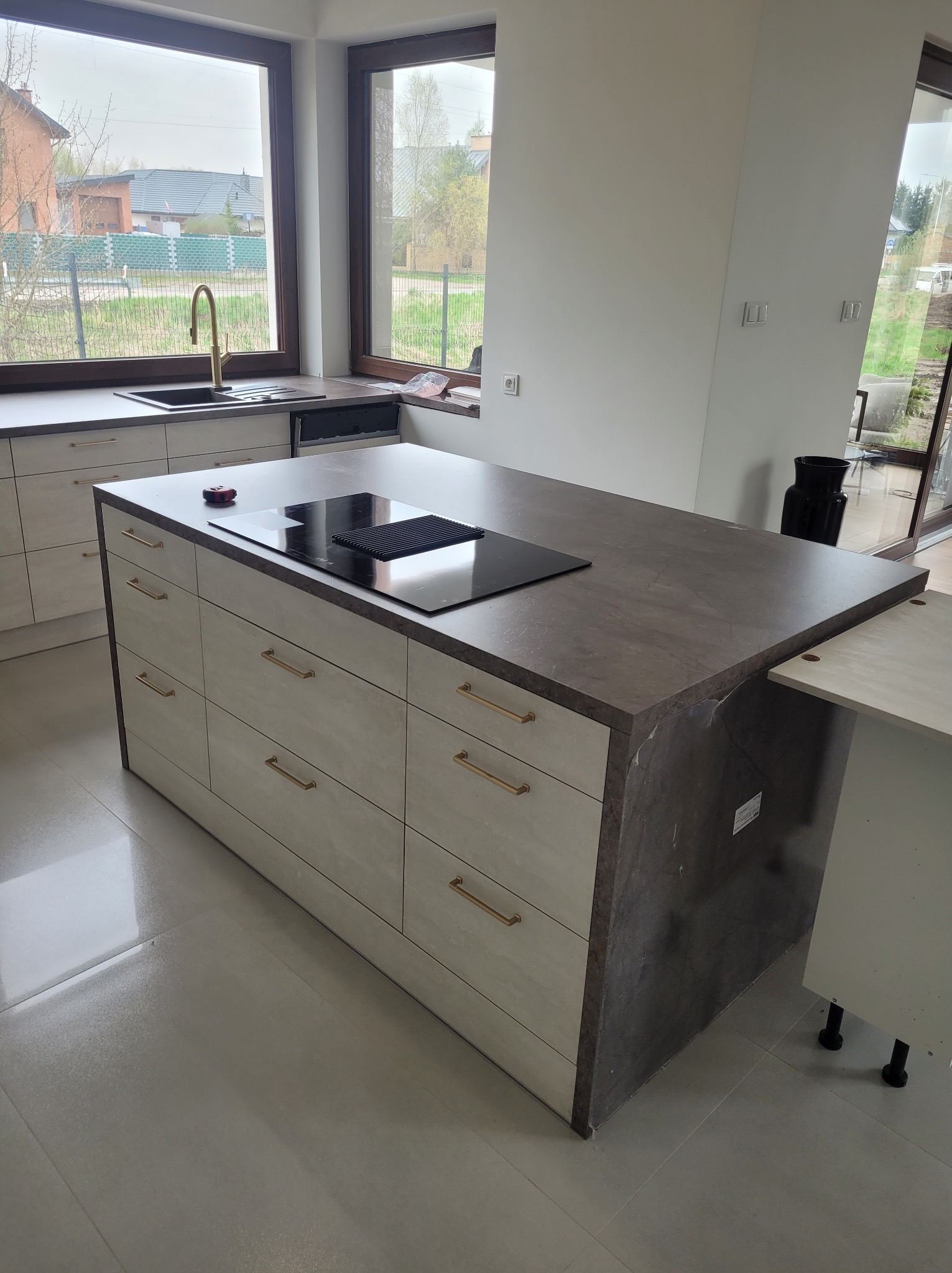 Wyspa kuchenna z szufladami i płytą indukcyjną, złote uchwyty, blat w odcieniach szarości, widok na okno i otoczenie domu.