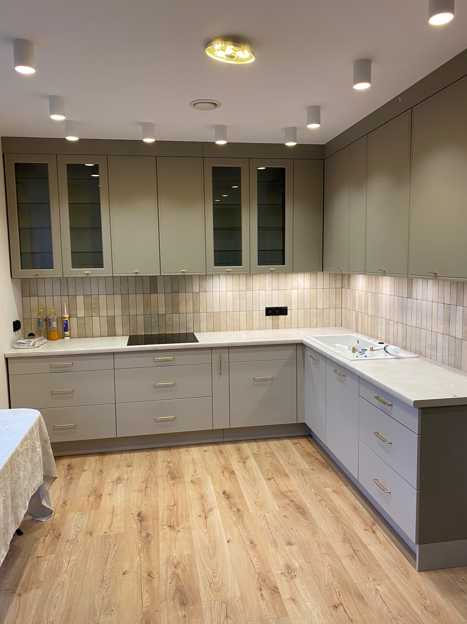 Jasna kuchnia z szarymi frontami, złotymi uchwytami i drewnianą podłogą. Widoczne górne szafki z przeszklonymi drzwiczkami oraz biały blat z płytą indukcyjną i zlewozmywakiem. Oświetlenie punktowe...