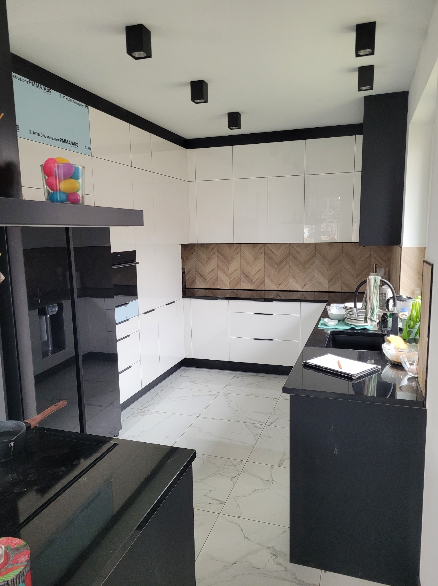 Nowoczesna kuchnia z białymi, błyszczącymi frontami szafek i czarnymi akcentami, parkiet ułożony w jodełkę na ścianie, marmurowe płytki na podłodze, czarne lampy sufitowe, szklanka z kolorowymi...