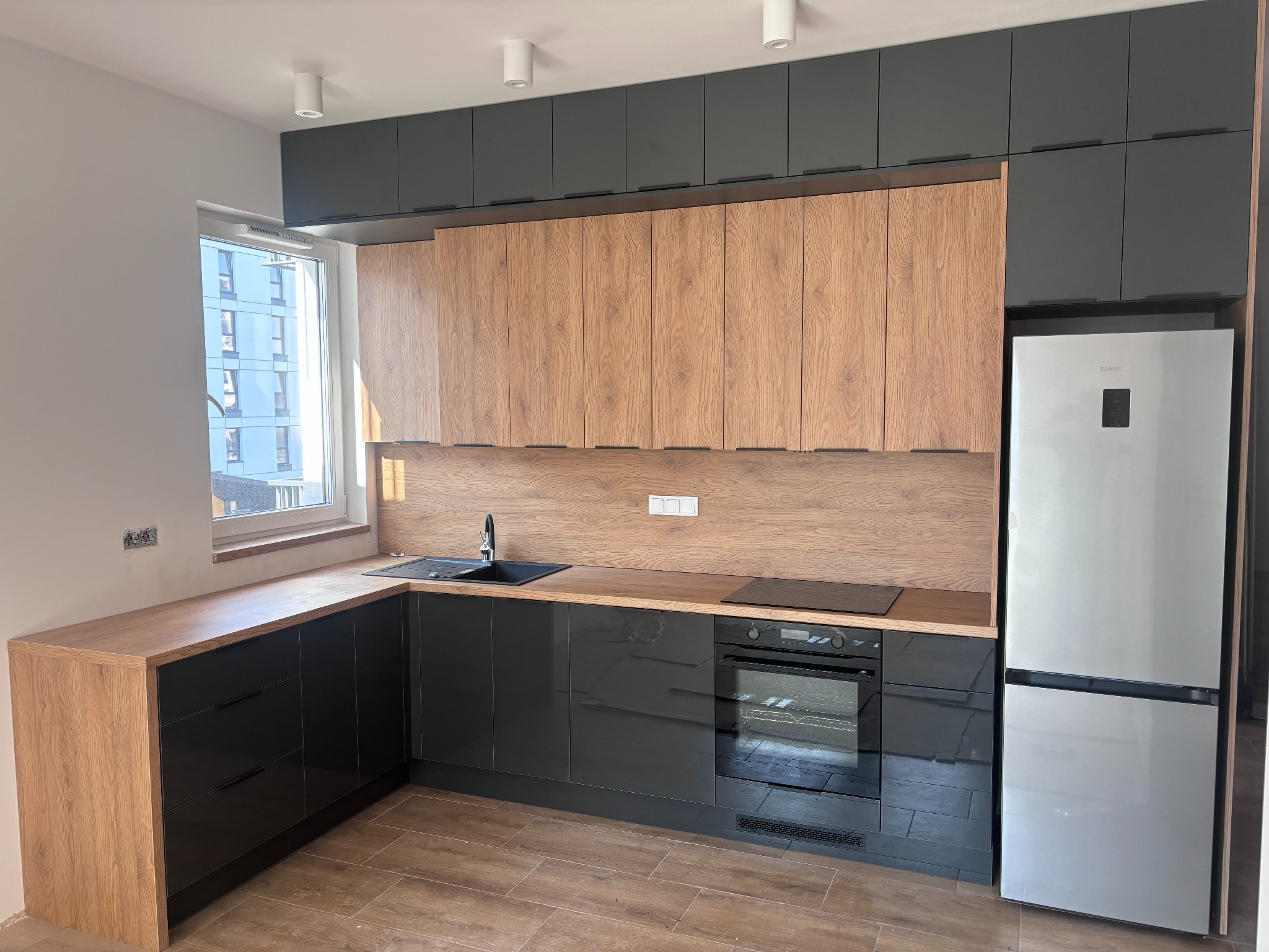 Nowoczesna kuchnia z drewnianymi szafkami górnymi i czarnymi szafkami dolnymi z połyskiem, czarnym zlewem i srebrną lodówką, oświetlona punktowymi lampami sufitowymi.