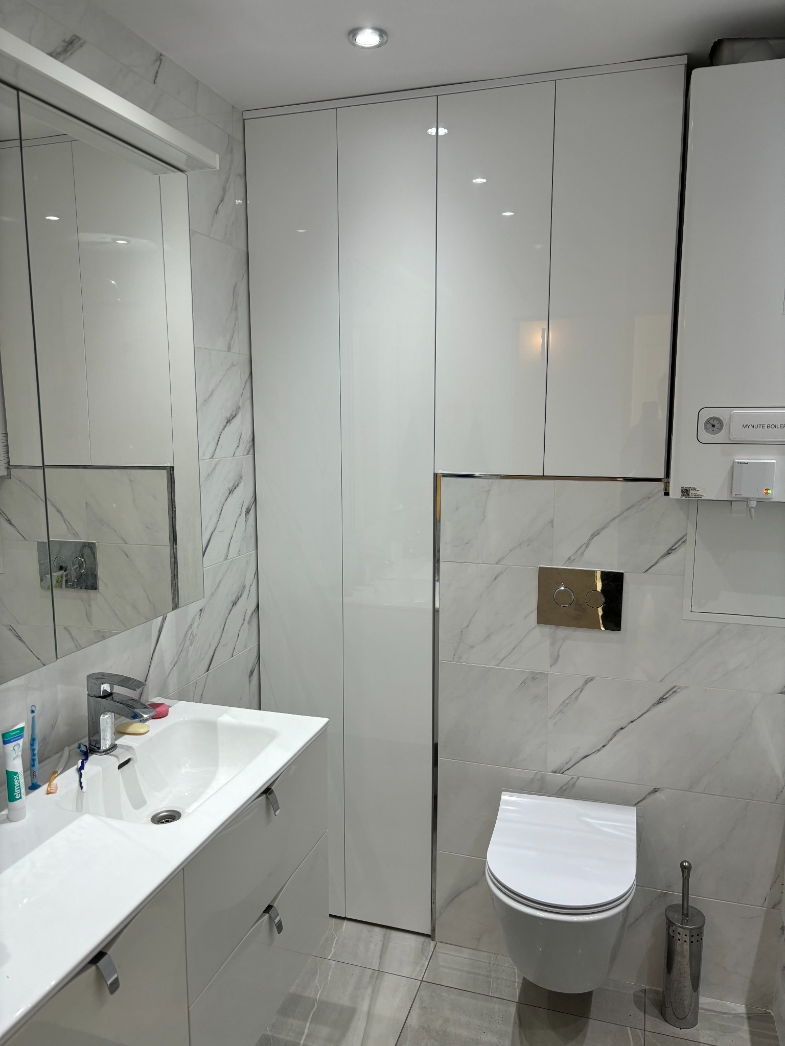 Nowoczesna, biała łazienka z marmurowymi płytkami, wiszącą toaletą i szafką pod umywalką, zintegrowana z wysoką szafą i wiszącym piecem gazowym.