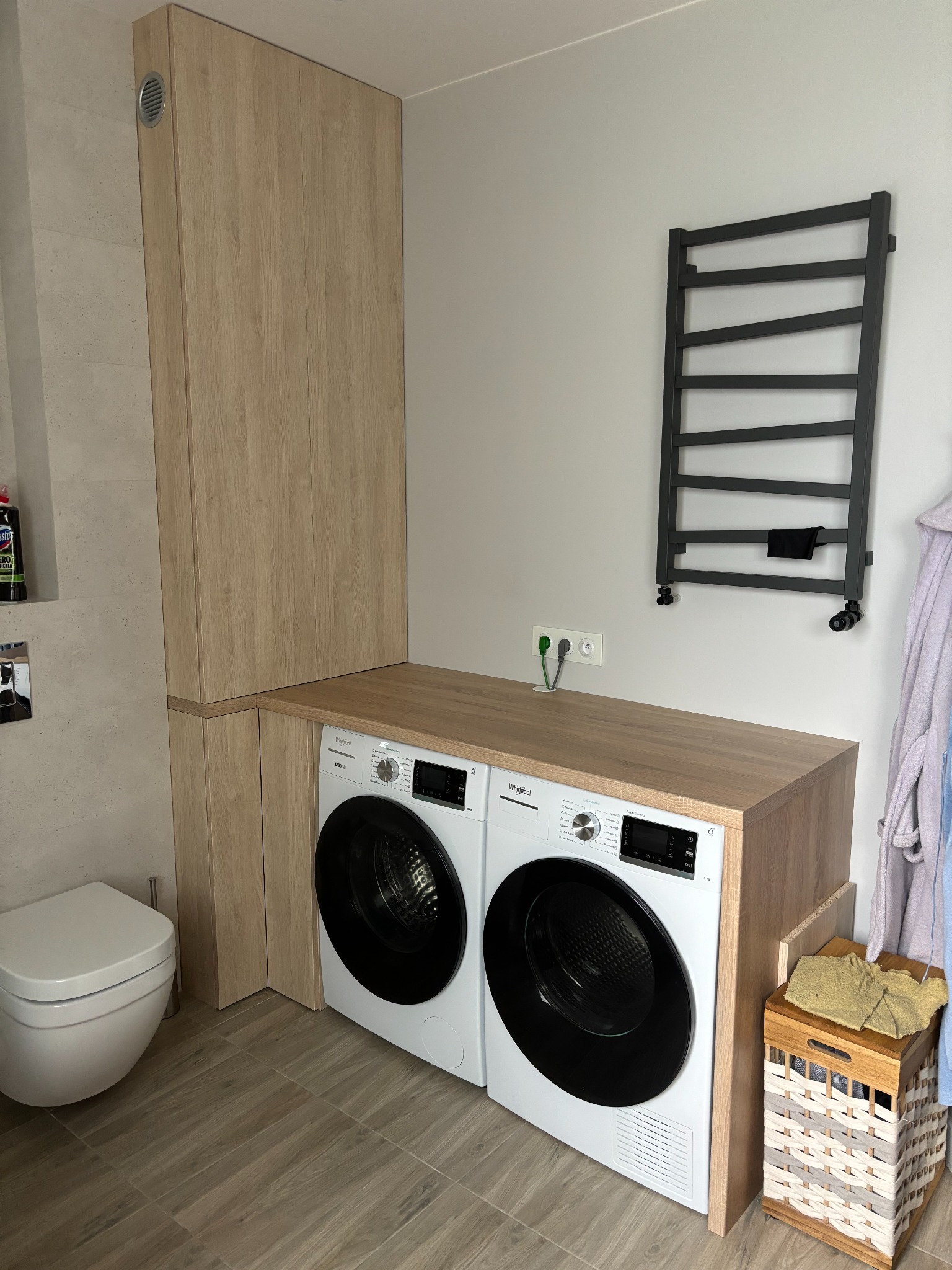 Zabudowa pralki i suszarki z szafką w jasnym drewnie w łazience z grzejnikiem drabinkowym i koszem na pranie.