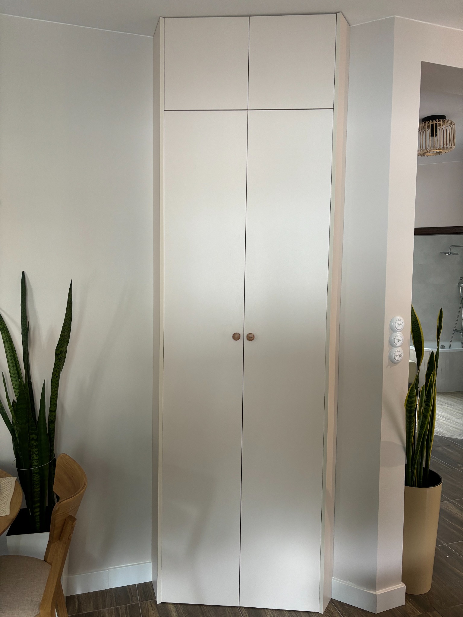 Wysoka, biała szafa z dwiema parami drzwi i okrągłymi, drewnianymi uchwytami, wkomponowana w korytarz, obok roślin doniczkowych i drewnianego krzesła. W tle widoczna łazienka z wanną i prysznicem.
