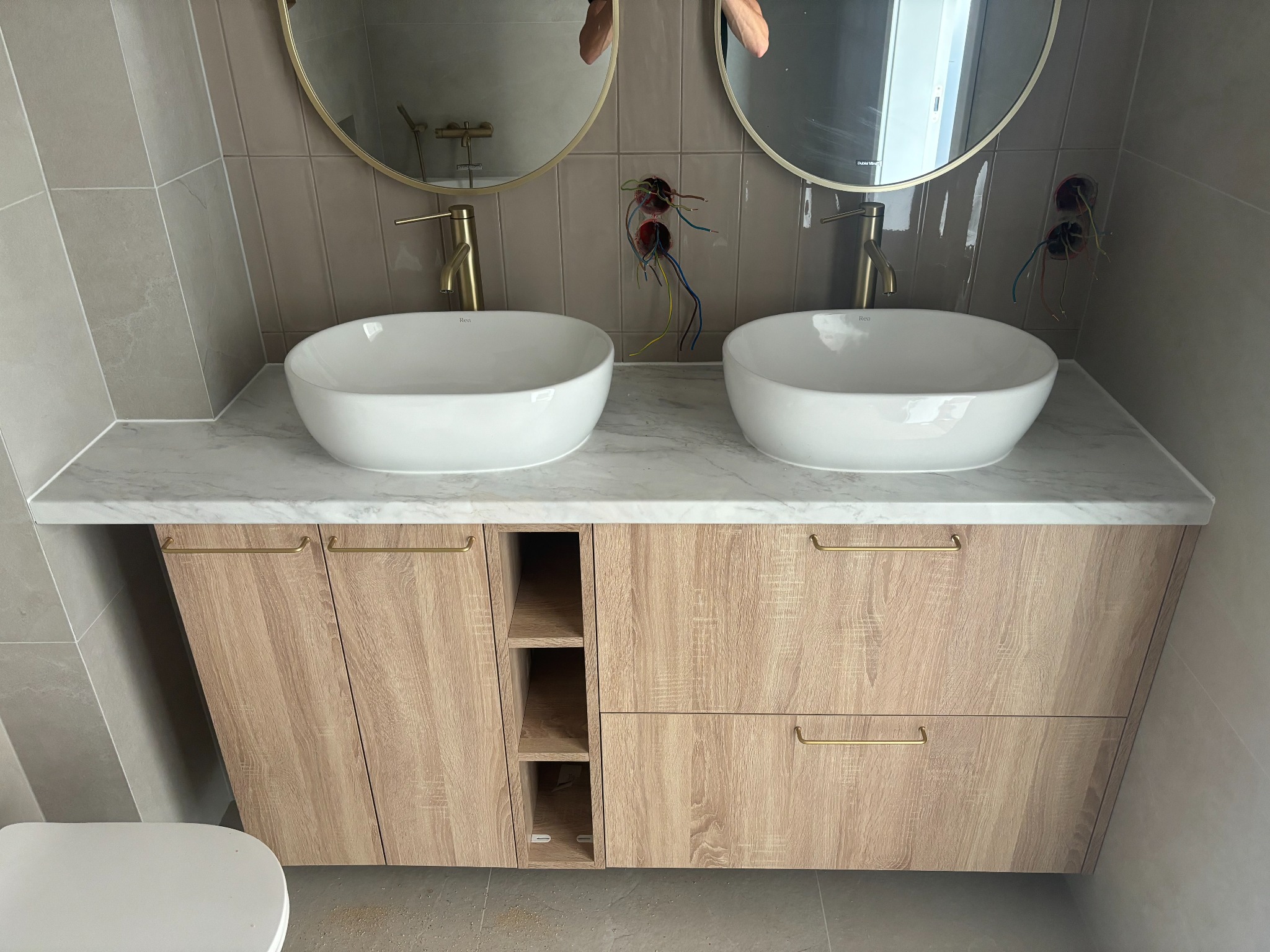 Podwójna umywalka nablatowa z marmurowym blatem i szafką z jasnego drewna, złote baterie i okrągłe lustra, widoczne niedokończone podłączenia elektryczne.