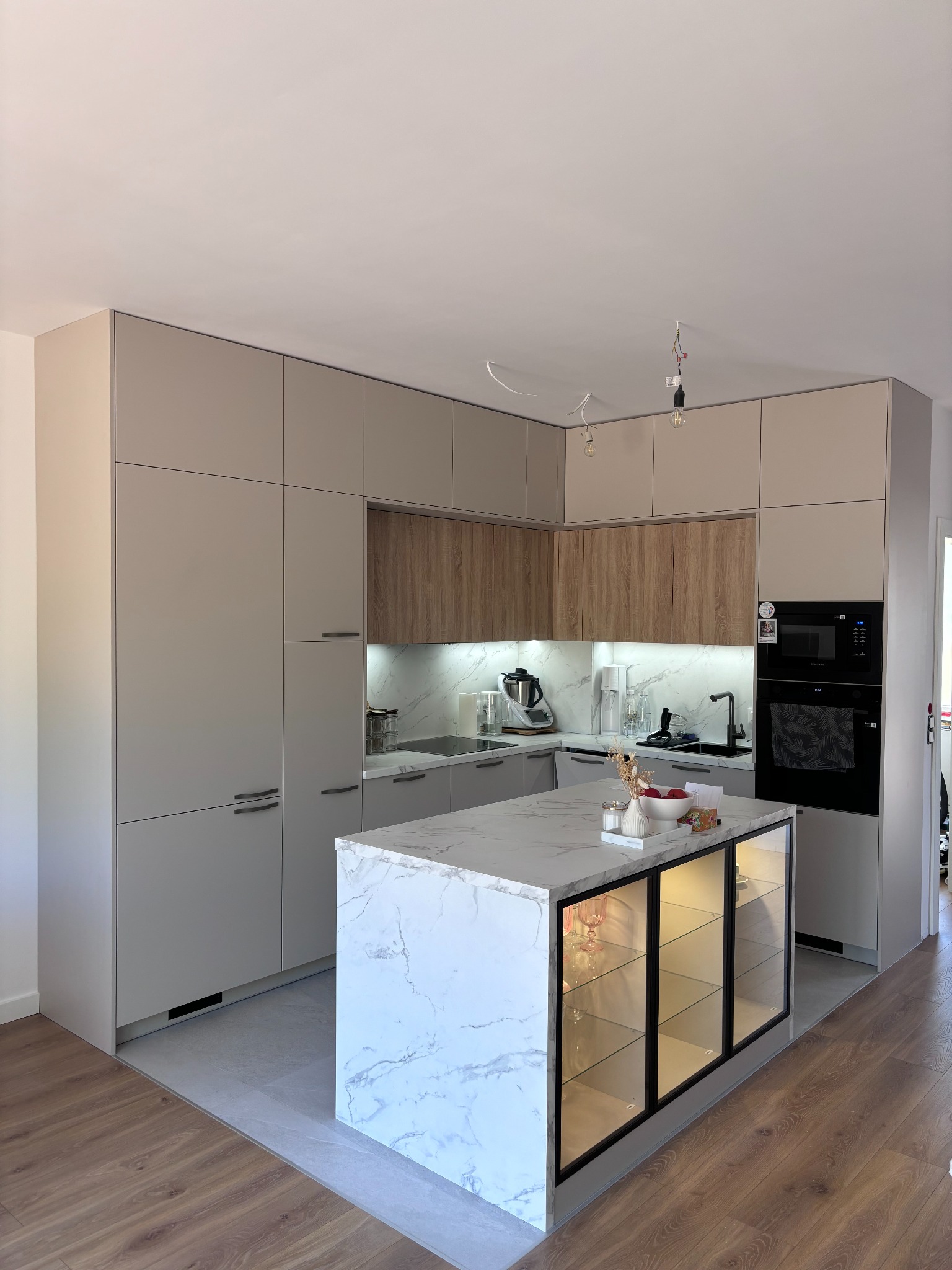 Nowoczesna kuchnia z beżowymi szafkami, drewnianymi akcentami i wyspą z blatem imitującym marmur, przeszklone szafki w wyspie podświetlone od wewnątrz, w tle widoczny sprzęt AGD