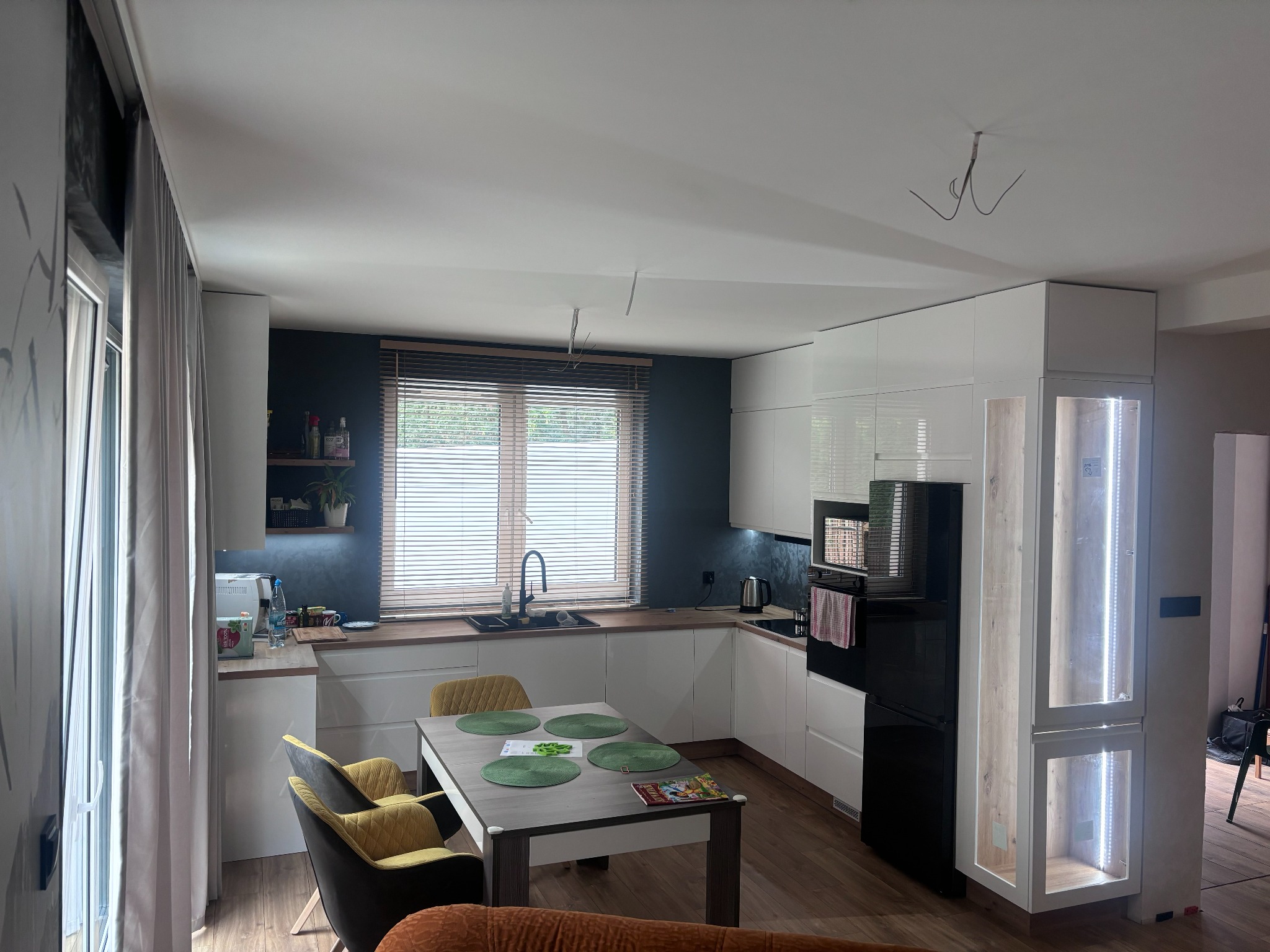 Jasna kuchnia z białymi frontami, drewnianym blatem i czarną ścianą w tle, okrągłe podkładki na stole i niedokończone oświetlenie sufitowe.