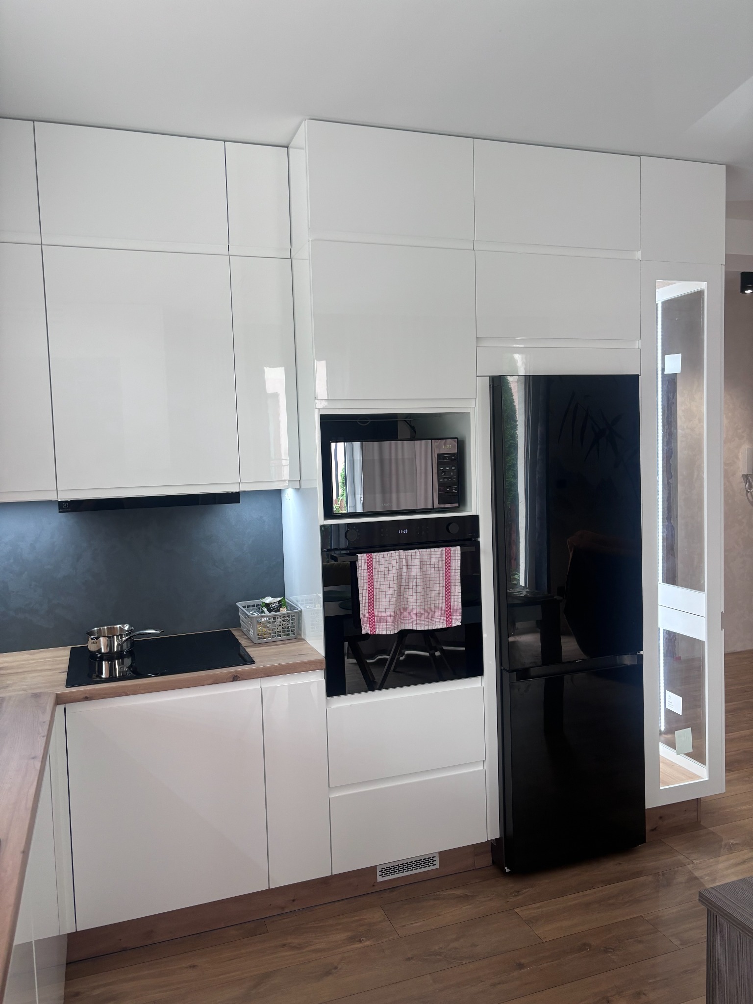 Nowoczesna kuchnia z białymi, lakierowanymi frontami, czarnym sprzętem AGD w zabudowie i drewnianym blatem, uzupełniona o płytę indukcyjną z garnkiem i koszyk z przyprawami.