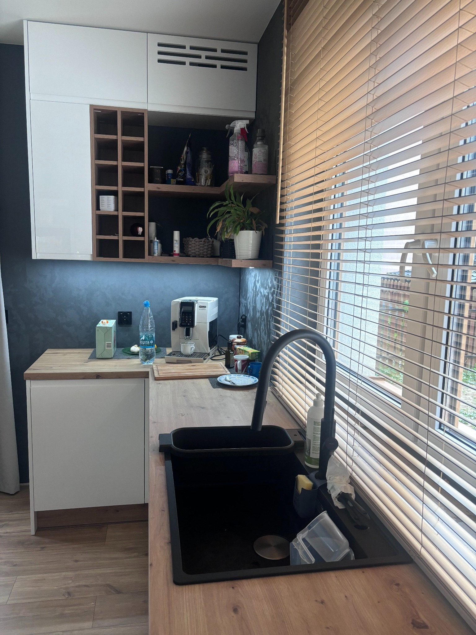 Fragment nowoczesnej kuchni z białymi szafkami, drewnianym blatem i czarnym zlewem pod oknem z żaluzjami. Na blacie ekspres do kawy, woda mineralna i akcesoria kuchenne.