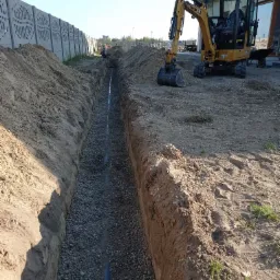 Wykopany rów z ułożoną rurą i obsypany żwirem, z żółtą koparką CAT na brzegu, w tle betonowy płot z ażurowym wzorem.