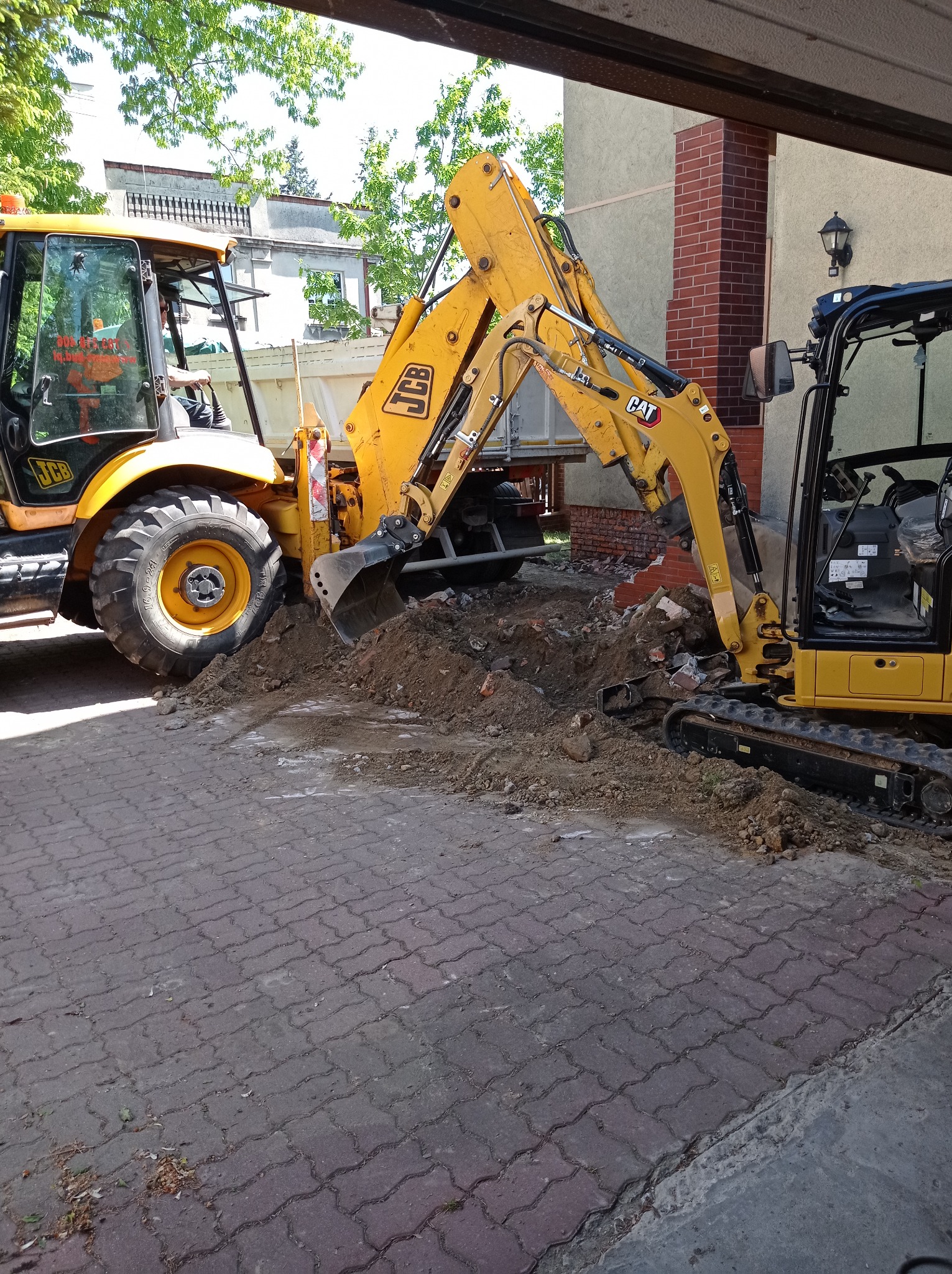 Żółta koparka JCB i minikoparka CAT usuwają gruz ceglany po wyburzeniu fragmentu ściany z czerwonej cegły na zewnątrz budynku.