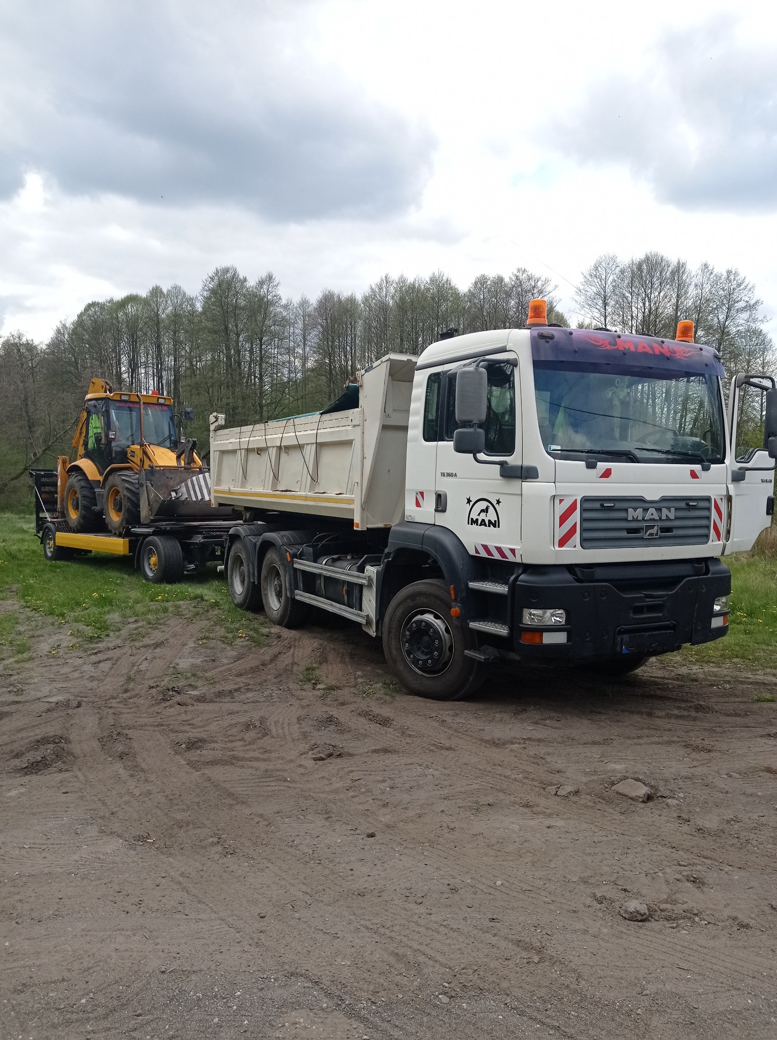 Ciężarówka MAN z otwartymi drzwiami kabiny i przyczepą z koparko-ładowarką JCB na błotnistym terenie w Mroczy.