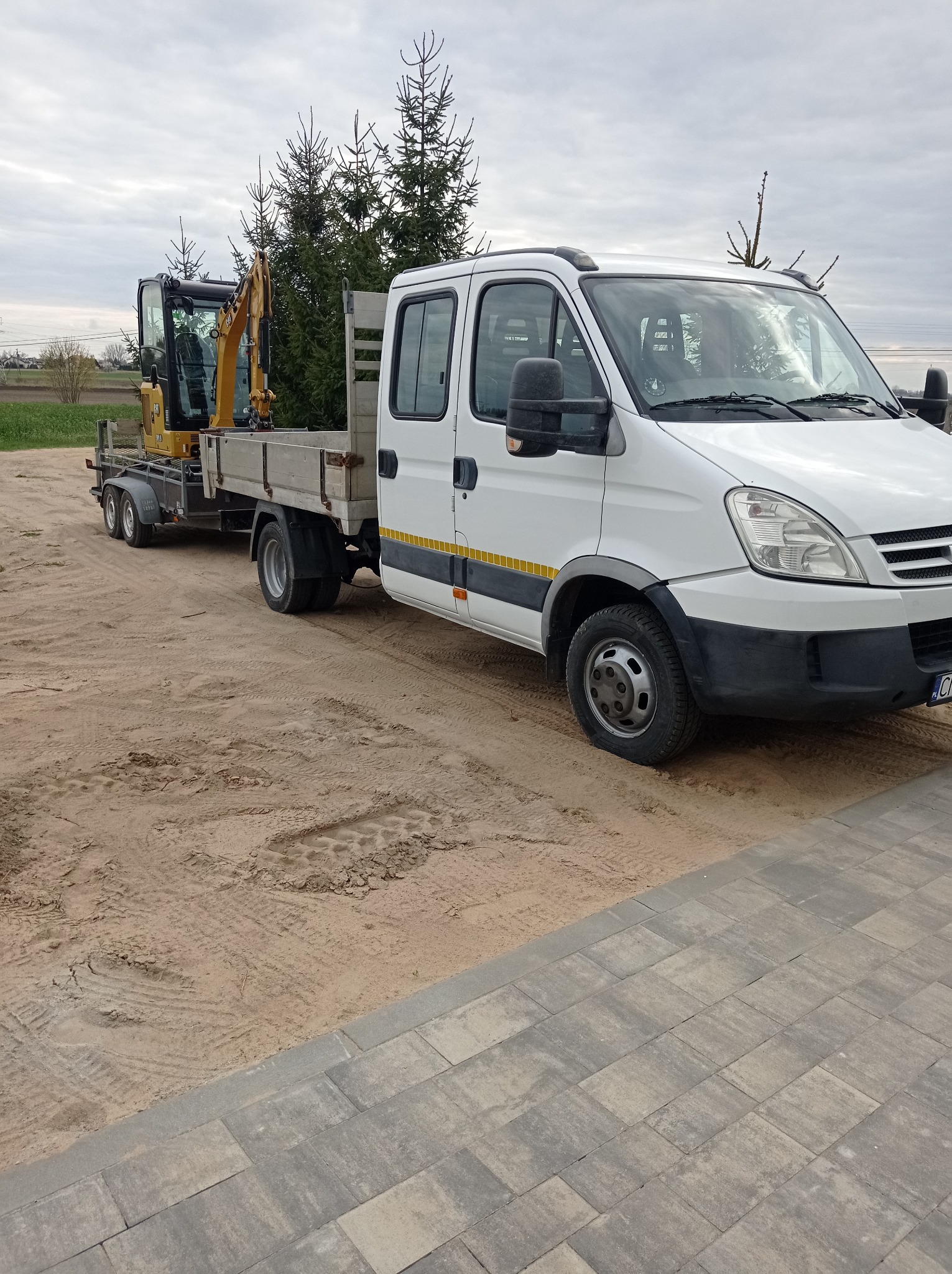 Biały samochód dostawczy z przyczepą, na której znajduje się mała koparka, zaparkowany na piaszczystym terenie obok nowej kostki brukowej.