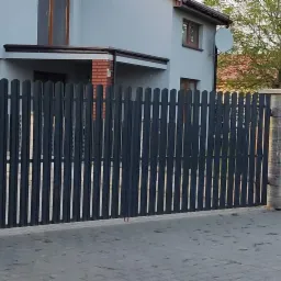 Nowoczesna, grafitowa brama wjazdowa z pionowych profili, osadzona na słupkach z szarej kostki łupanej, na tle jasnego domu z elementami cegły.