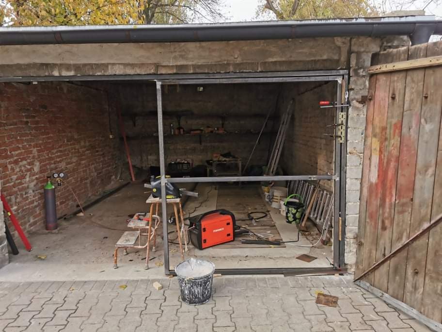 Metalowa rama bramy garażowej w trakcie montażu, widoczne narzędzia spawalnicze, butle z gazem, garaż z ceglanymi ścianami w tle.