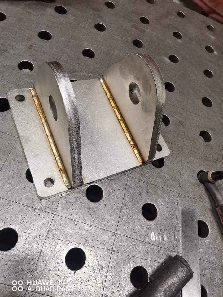 Metalowy element konstrukcyjny, spawany na stole spawalniczym z widocznymi śladami spawania i otworami montażowymi.