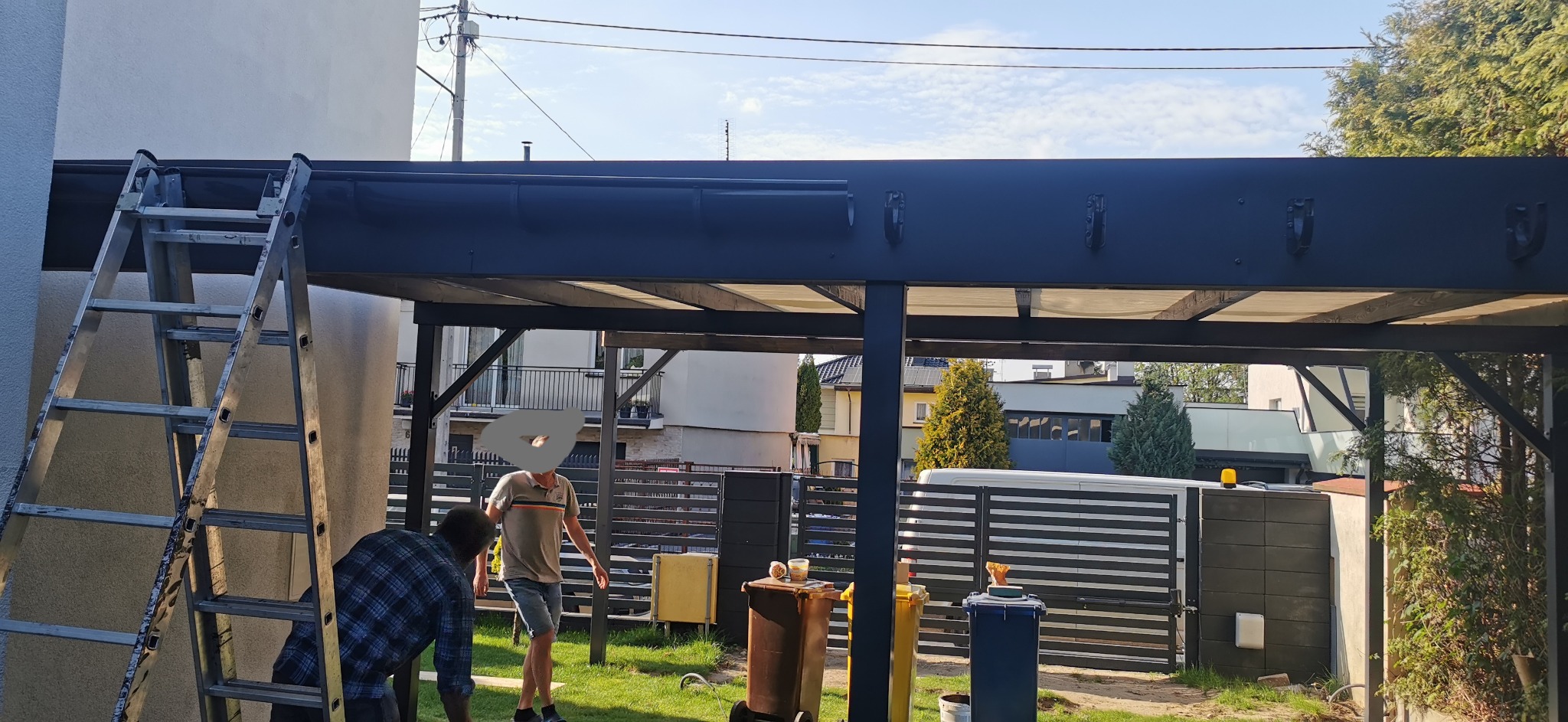 Montaż zadaszenia tarasu w Księstwie: widok na konstrukcję z czarnych metalowych profili, drabina oparta o ścianę budynku, pracownicy w trakcie instalacji, w tle zabudowa jednorodzinna i ogrodzenie...
