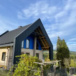 Dom w trakcie prac dekarskich, widoczna czarna blacha na dachu i niebieska membrana, częściowo odsłonięta konstrukcja dachu, rusztowanie i drabina przy ścianie, widok na zielone wzgórza w tle.