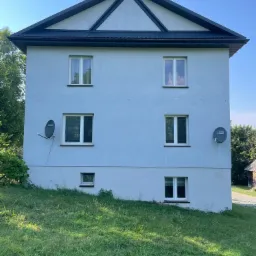 Dwupiętrowy dom z szarym dachem z blachy trapezowej i antenami satelitarnymi na elewacji, widziany z poziomu trawnika.