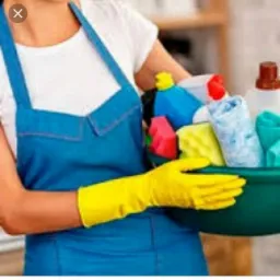 Osoba w niebieskim fartuchu i żółtych rękawicach trzyma zielony pojemnik wypełniony środkami czystości i ściereczkami, przygotowana do pracy.