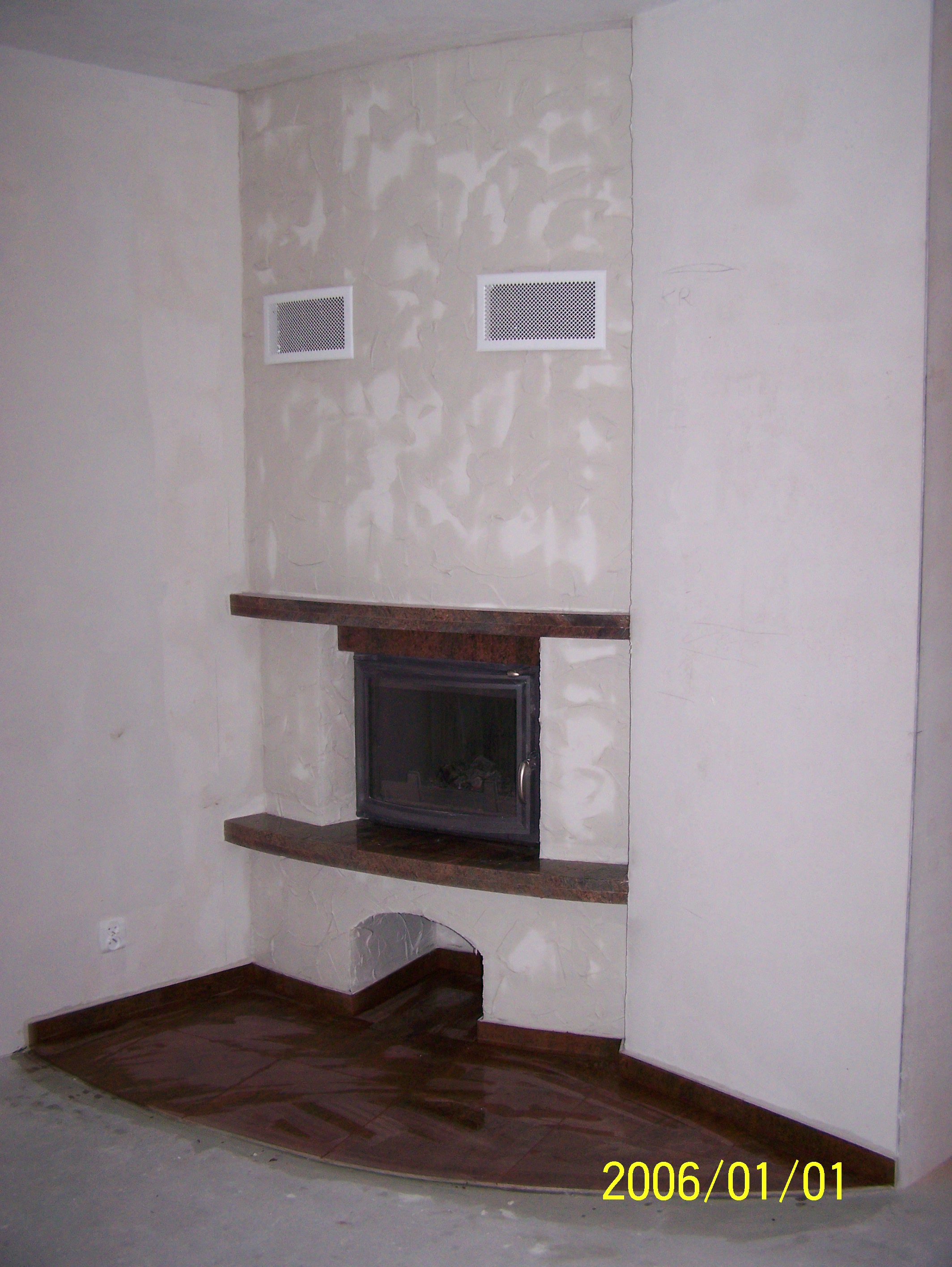 Wnętrze pokoju z niedokończonym kominkiem z kamiennymi półkami, widoczne kratki wentylacyjne w ścianie i wykończona podłoga, ściany w trakcie wykańczania.