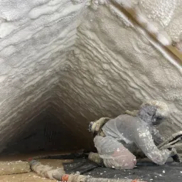 Pracownik w kombinezonie ochronnym aplikuje piankę izolacyjną w trudno dostępnym poddaszu, widoczne drewniane elementy konstrukcyjne i przewody robocze.