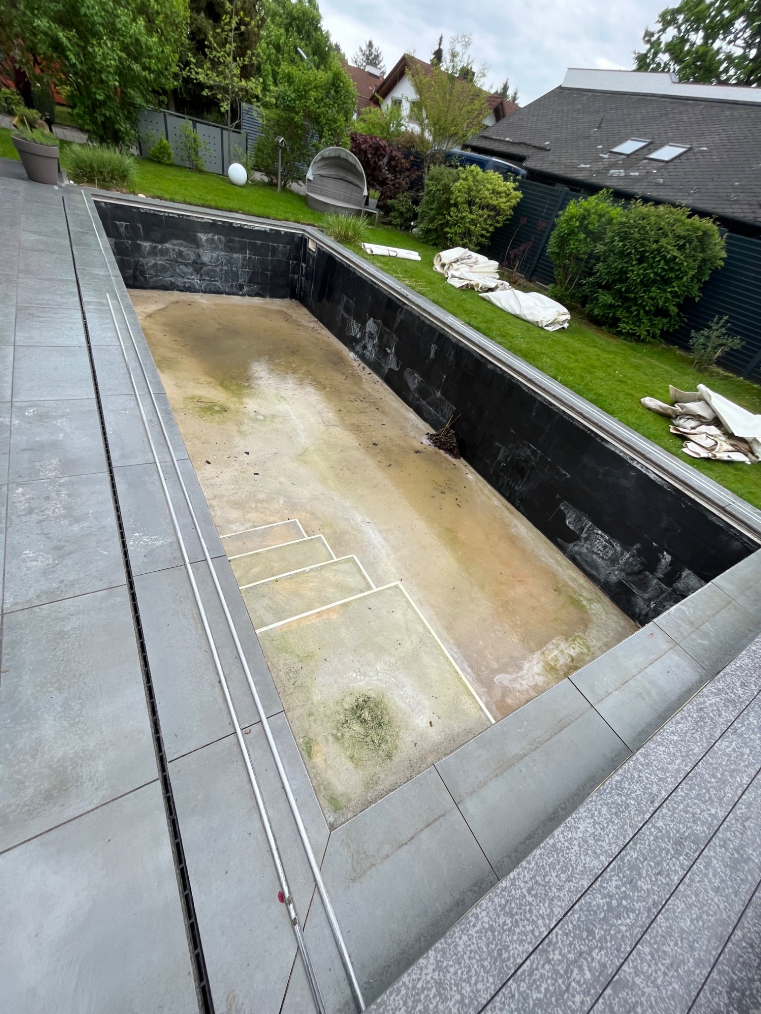 Opróżniony, brudny basen ogrodowy z widocznymi schodami i czarną izolacją ścian, otoczony szarymi płytkami, z zielonym trawnikiem i elementami wyposażenia ogrodu w tle.