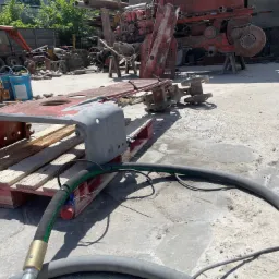 Część maszyny na palecie, przygotowana do obróbki, z widocznymi śladami rdzy i starej farby, obok wąż do piaskowania. W tle inne elementy maszyn i sprzęt budowlany.