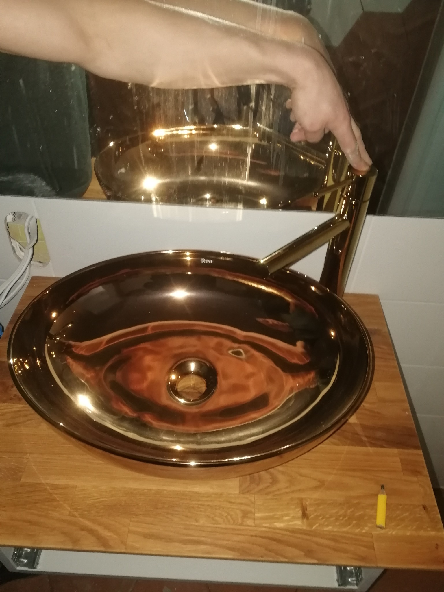 Błyszcząca, złota umywalka nablatowa z baterią, zamontowana na drewnianym blacie w łazience z lustrem. Widoczna dłoń osoby regulującej baterię.