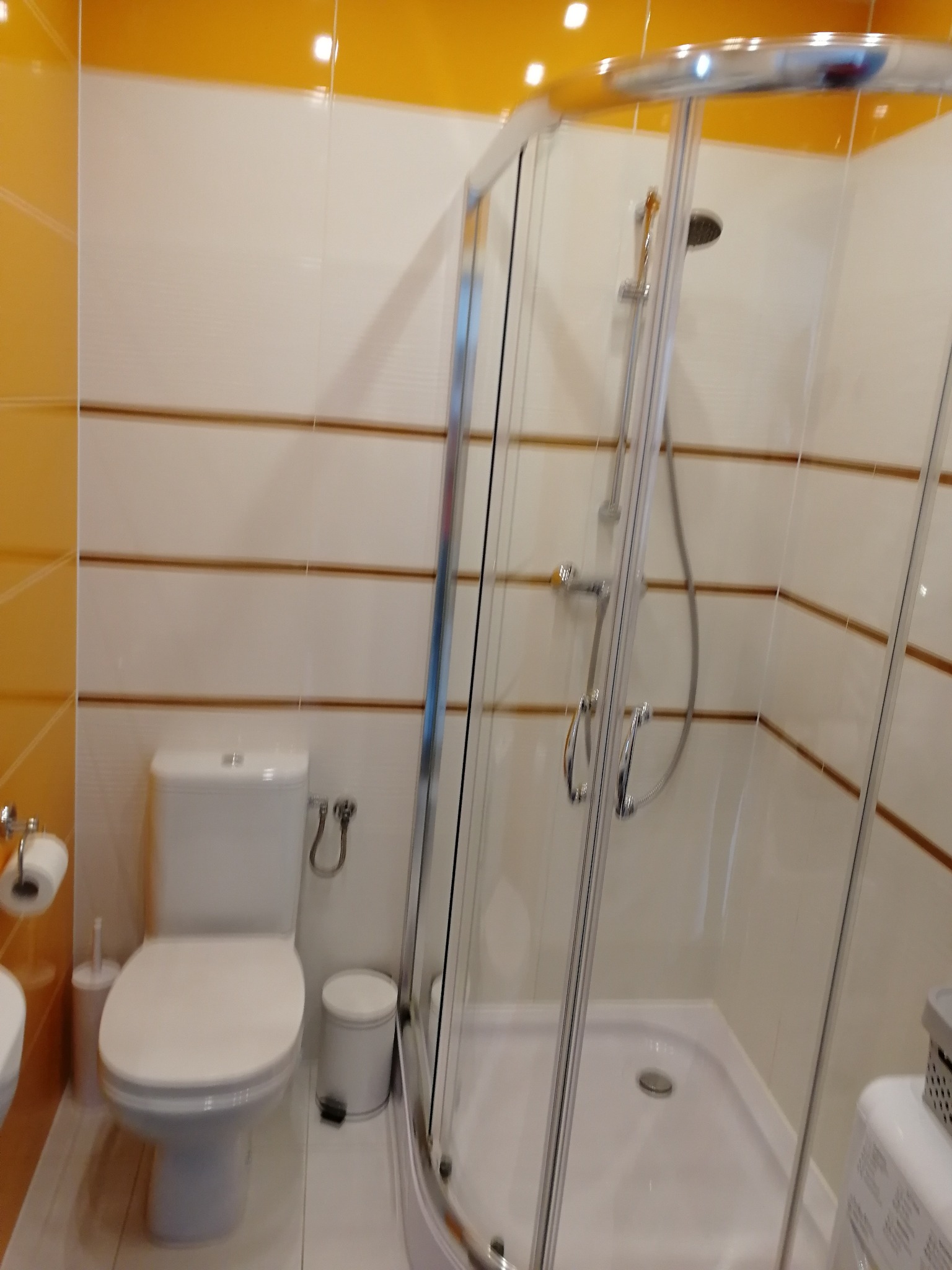 Jasna łazienka z żółtymi akcentami, kabina prysznicowa z chromowanymi profilami i biała toaleta z pojemnikiem na szczotkę, widoczne płytki z poziomymi paskami.