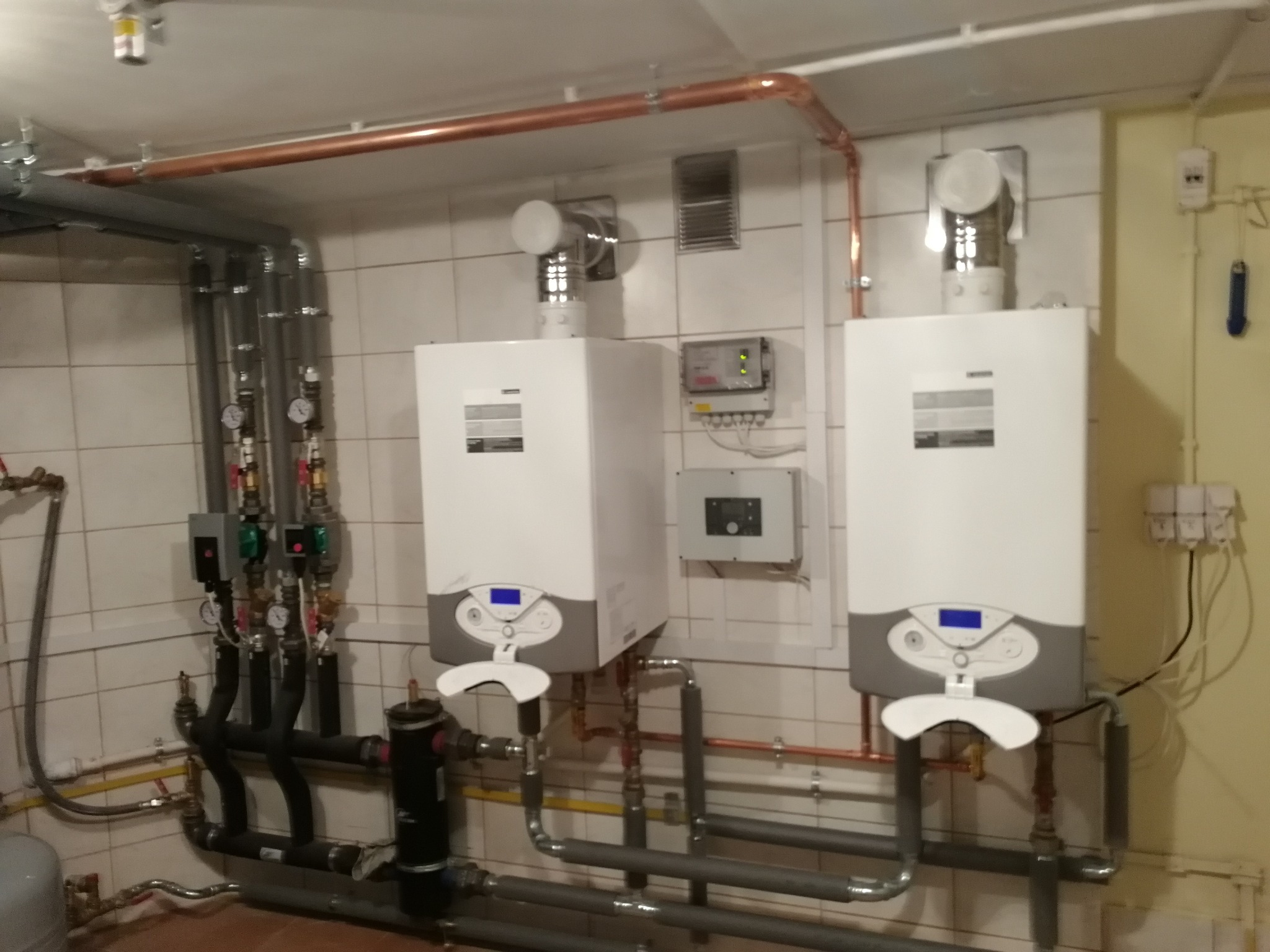 Dwa białe kotły grzewcze zamontowane na ścianie z beżowych płytek w Bydgoszczy, widoczne rury miedziane i szare, manometry oraz instalacja elektryczna.