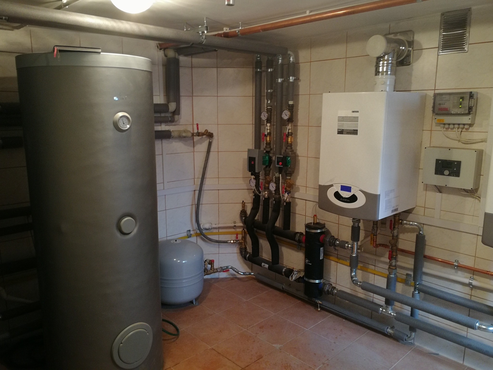 Kotłownia w beżowych płytkach z dużym szarym zasobnikiem ciepłej wody, białym piecem gazowym, szarym zbiornikiem wyrównawczym i licznymi rurami oraz zaworami.