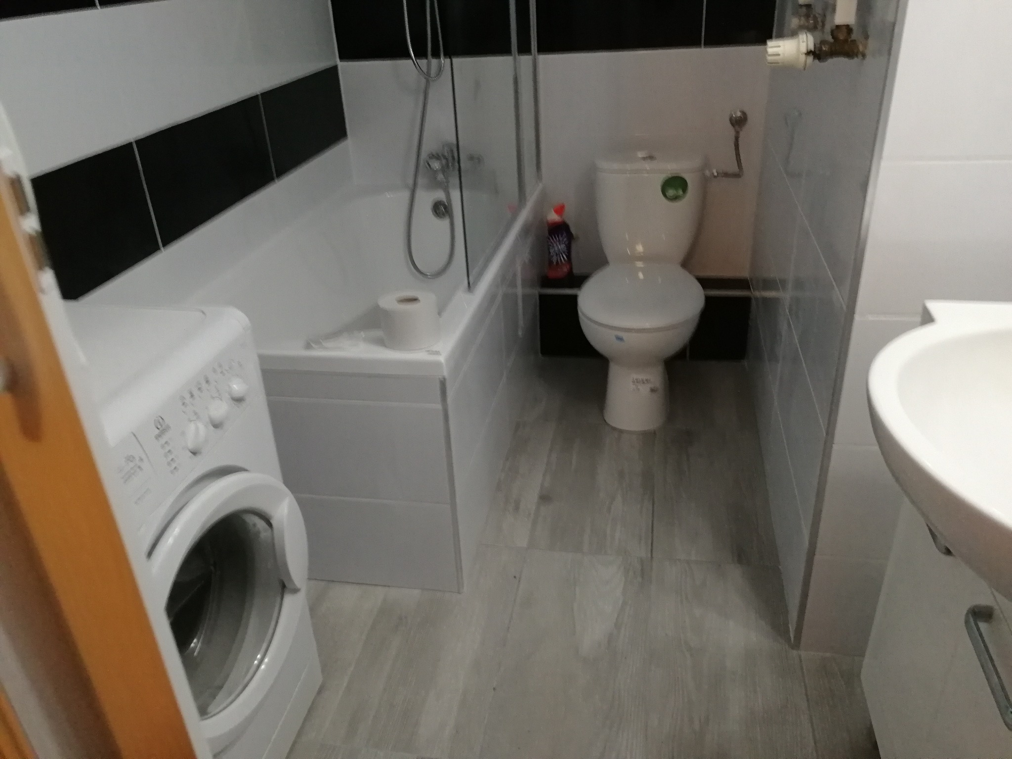 Wnętrze łazienki po remoncie: wanna z parawanem, toaleta, umywalka z szafką i pralka w odcieniach bieli i szarości, z akcentami czarnych płytek.