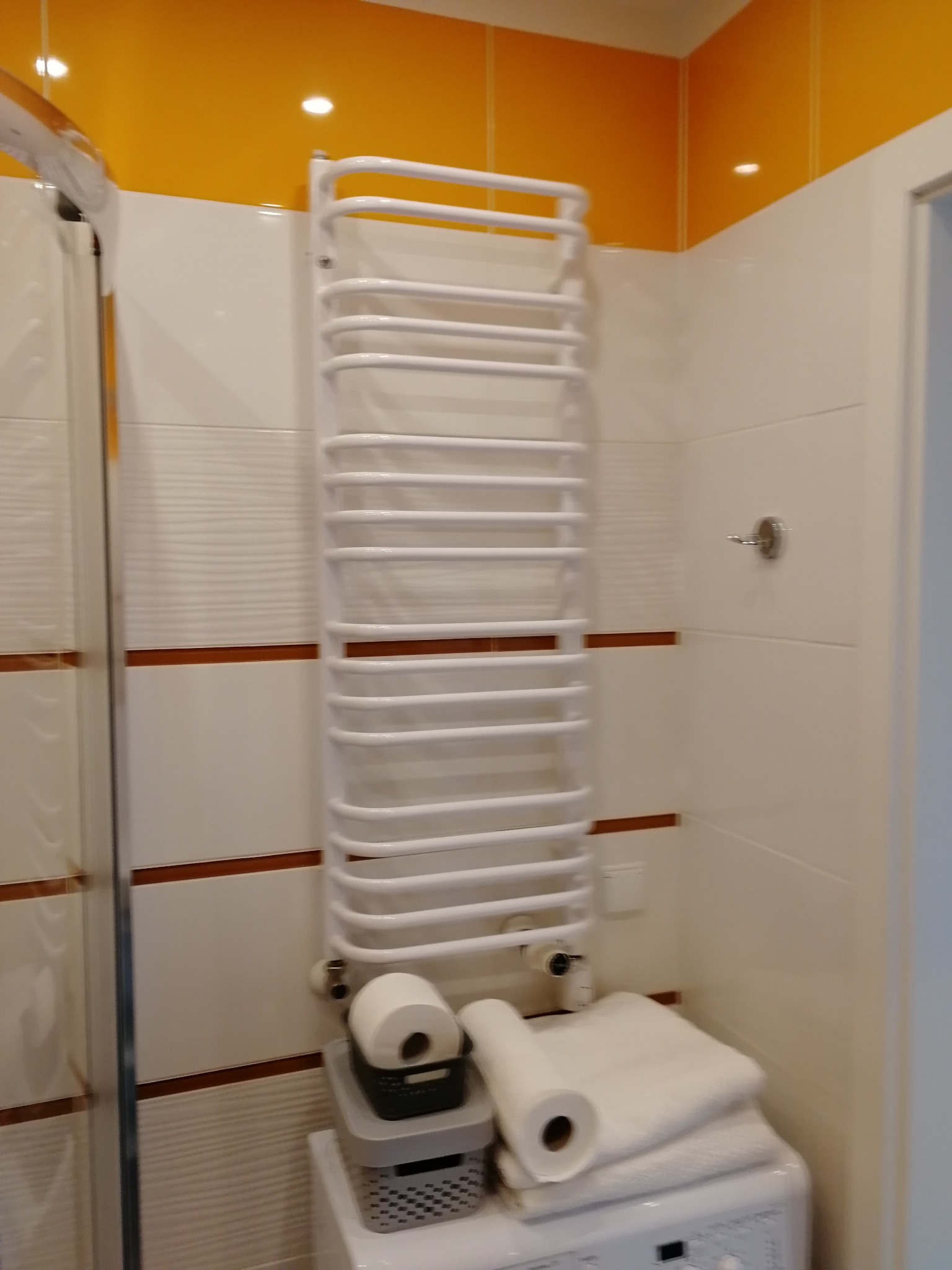 Biały grzejnik drabinkowy łazienkowy na ścianie z beżowymi i pomarańczowymi płytkami, z rolką papieru toaletowego w koszyku i ręcznikami na pralce.