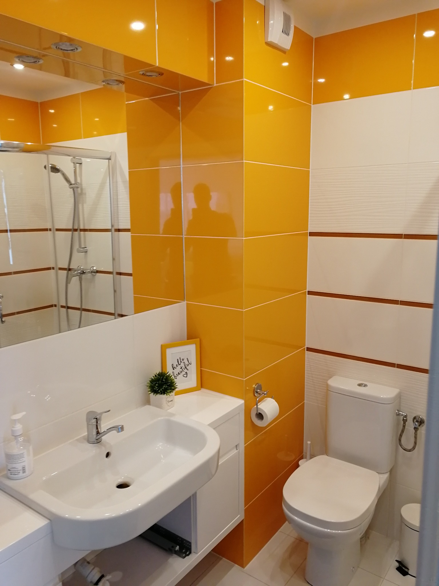 Jasna łazienka z żółtymi akcentami: biała umywalka z baterią, toaleta, kabina prysznicowa odbijająca się w lustrze, wentylator ścienny i dekoracyjna ramka ze sloganem.