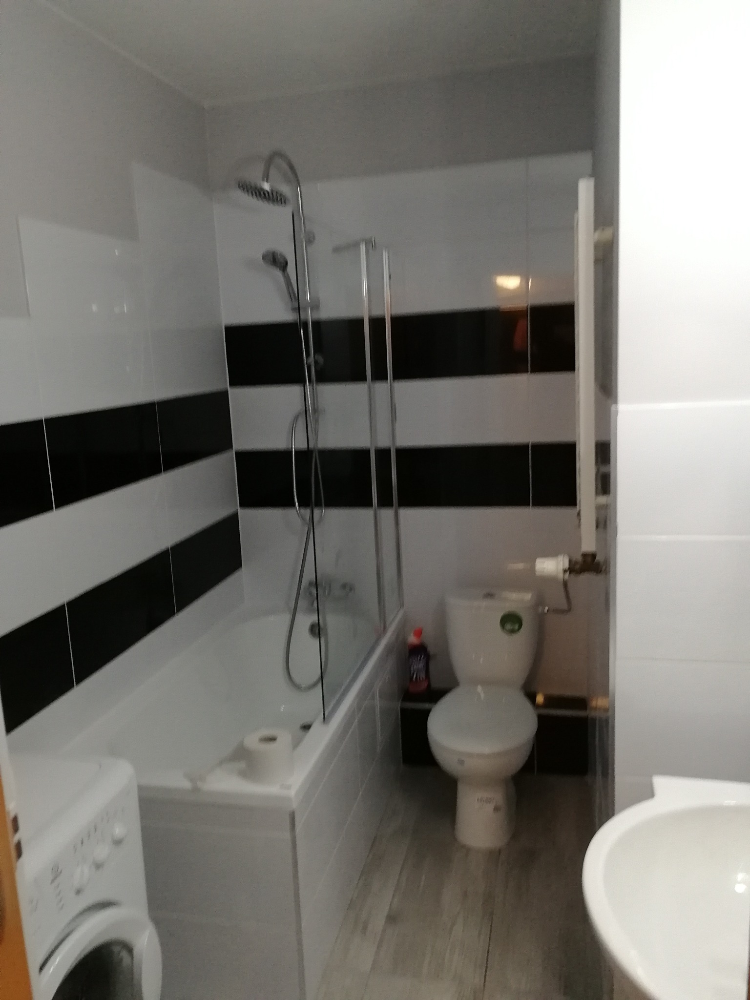 Wnętrze łazienki z wanną ze szklanym parawanem, toaletą, umywalką i pralką. Ściany wyłożone białymi płytkami z poziomymi czarnymi pasami. Na wannie rolka papieru toaletowego.