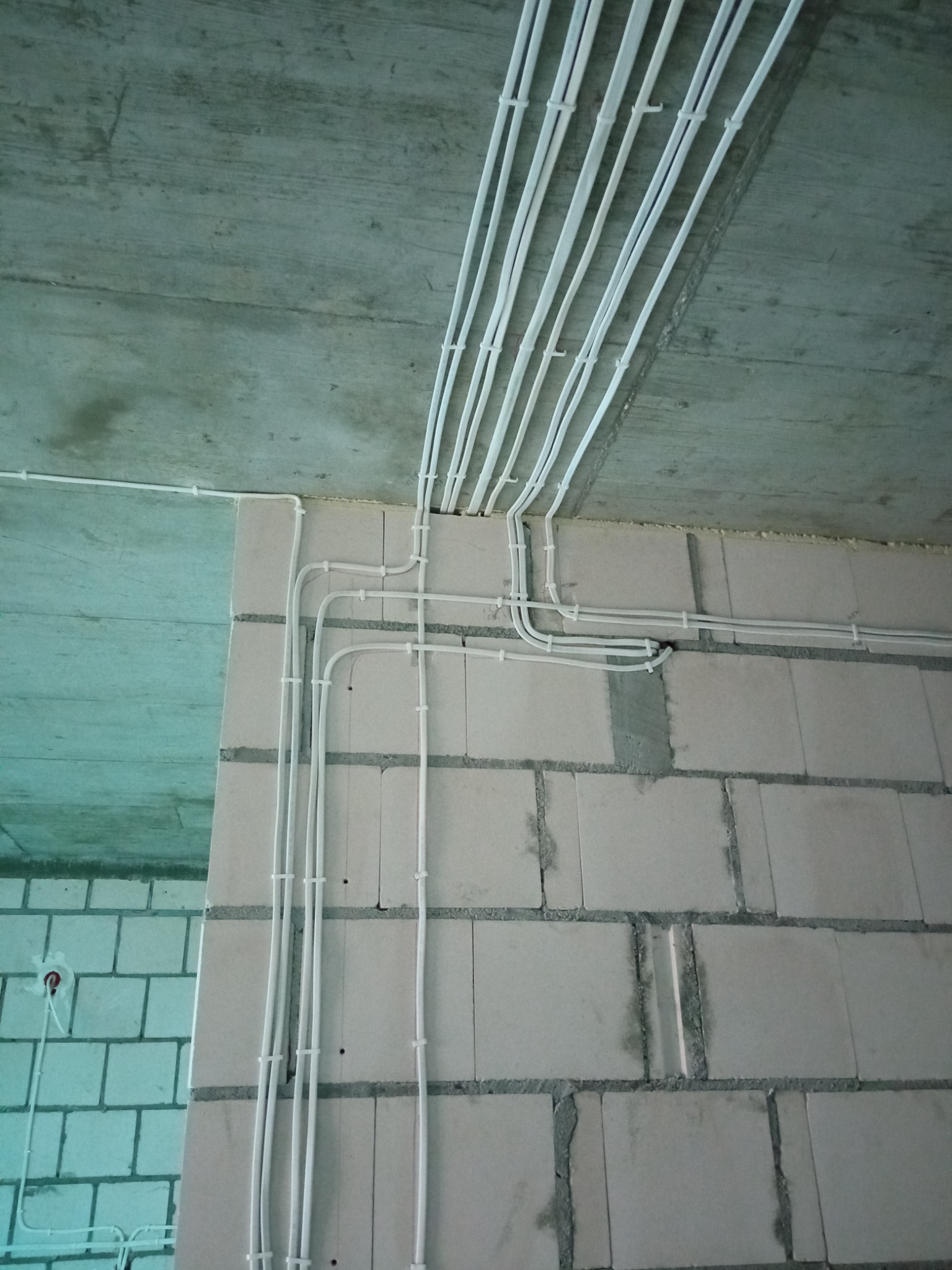 Białe przewody elektryczne prowadzone po powierzchni surowego sufitu i ściany z betonu komórkowego, zabezpieczone uchwytami, widoczne puszki elektryczne.