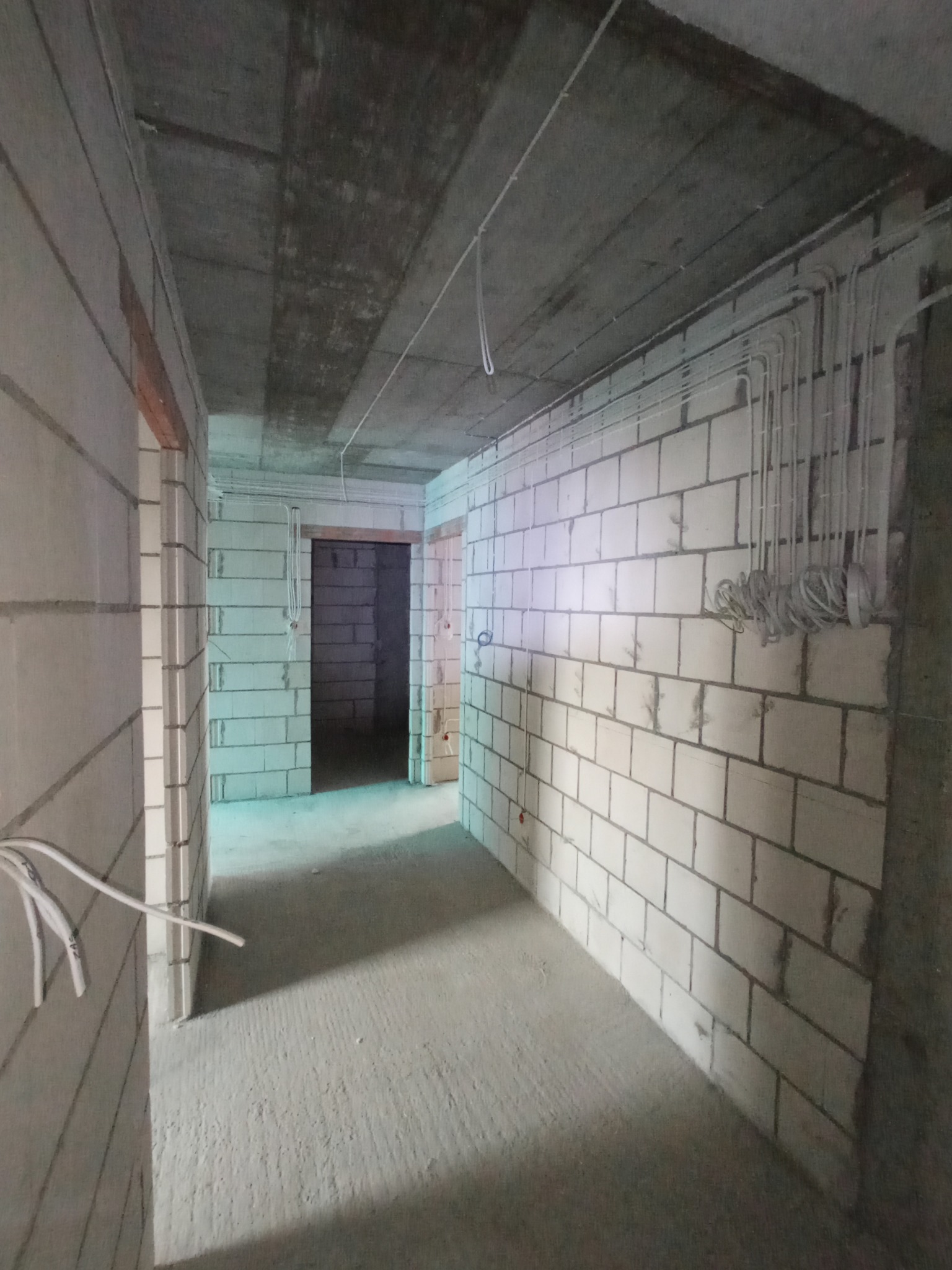 Surowy stan budynku w trakcie remontu, widoczne poprowadzone przewody elektryczne pod sufitem i wzdłuż ściany z białych bloczków, gołe ściany i betonowa podłoga.