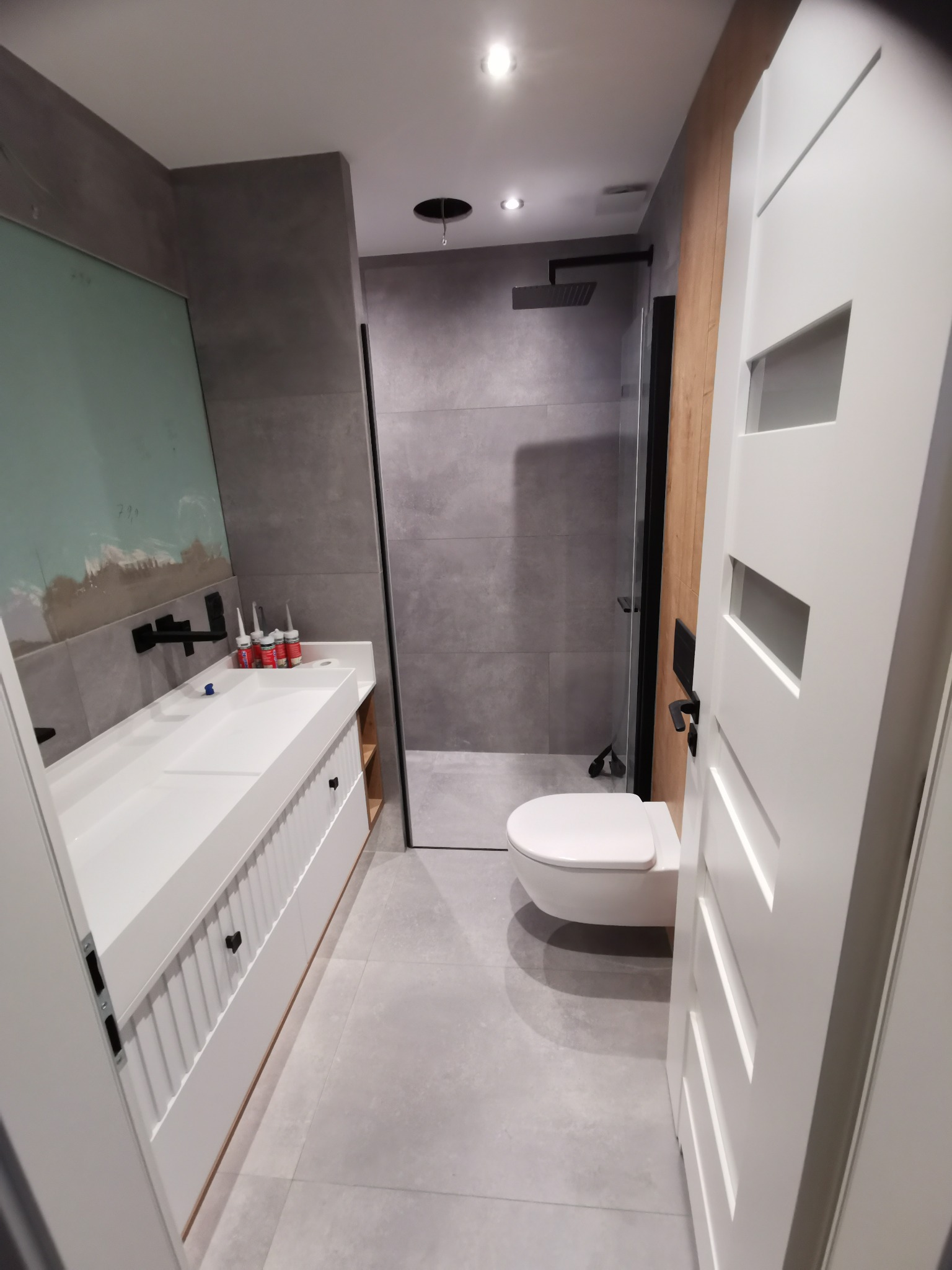 Nowoczesna łazienka z szarą płytką, białą umywalką nablatową, czarną armaturą i kabiną prysznicową typu walk-in, widoczne tuby silikonu przy umywalce.